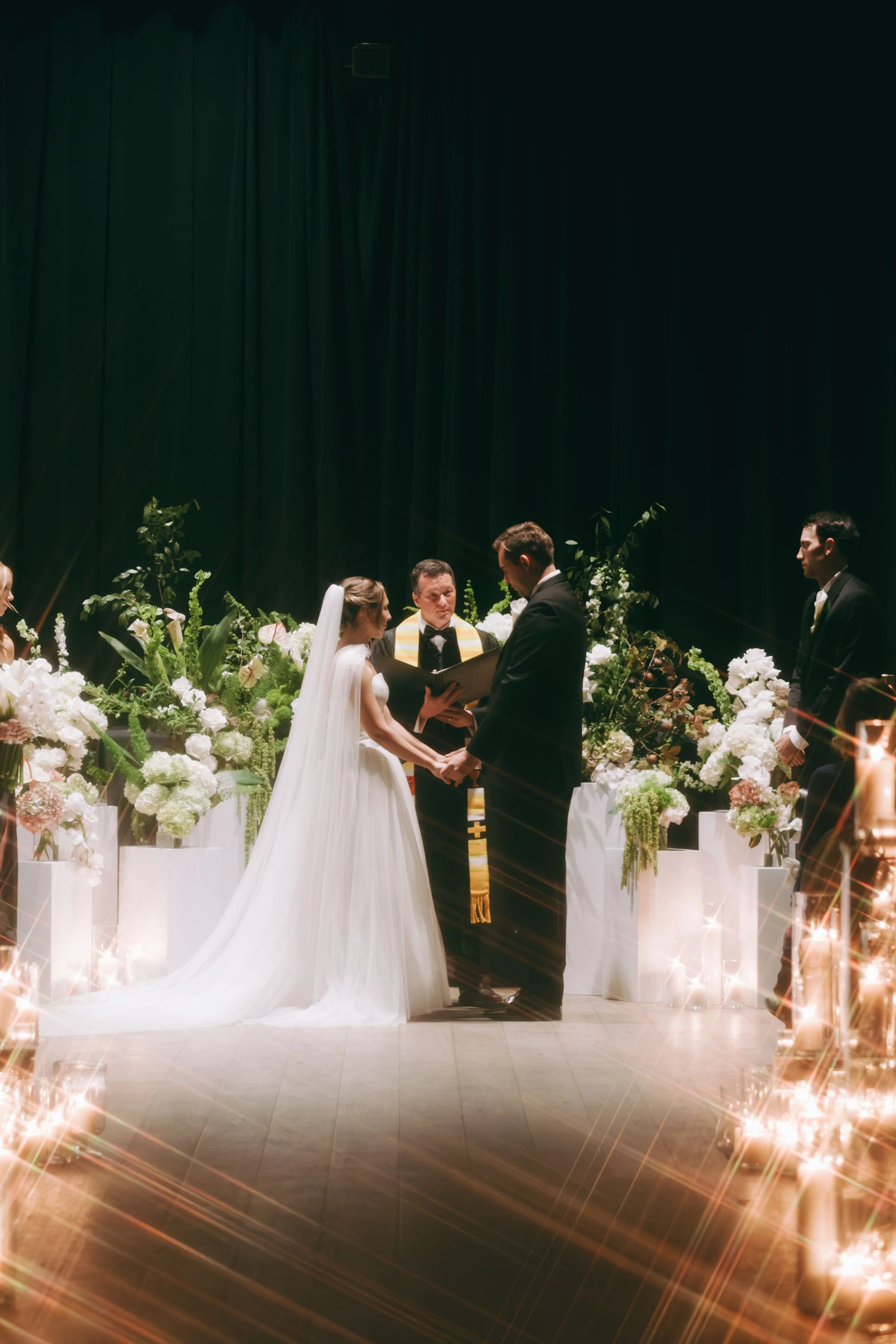 A wedding ceremony with a bride and groom holding hands, facing each other in front of an officiant, surrounded by white flowers and candles, with a green curtain backdrop.