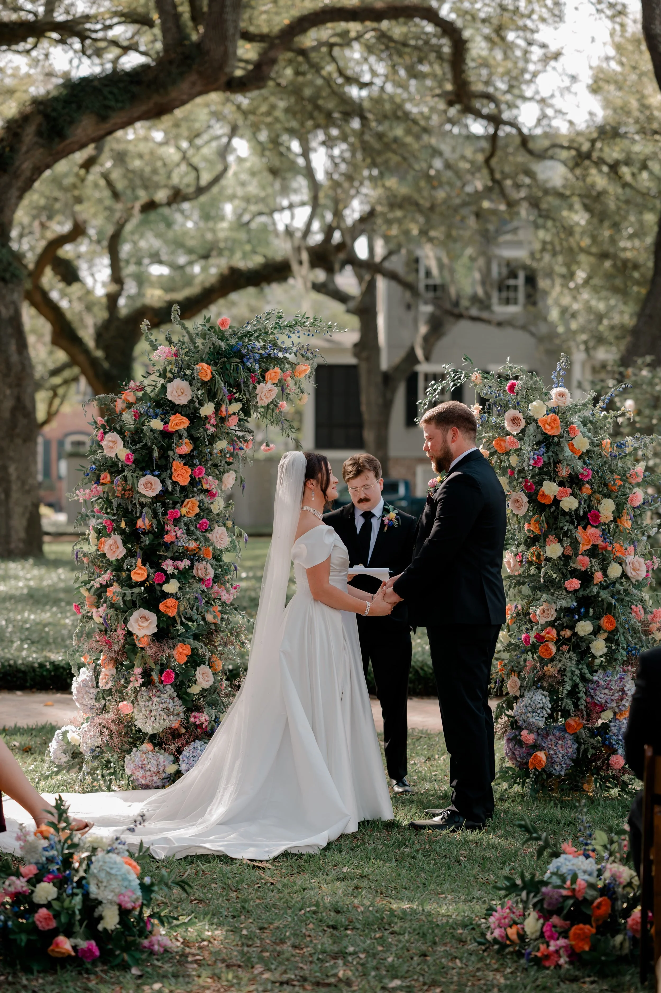 A bride and groom exchange vows under a floral arch during an outdoor wedding ceremony, surrounded by trees and floral arrangements.