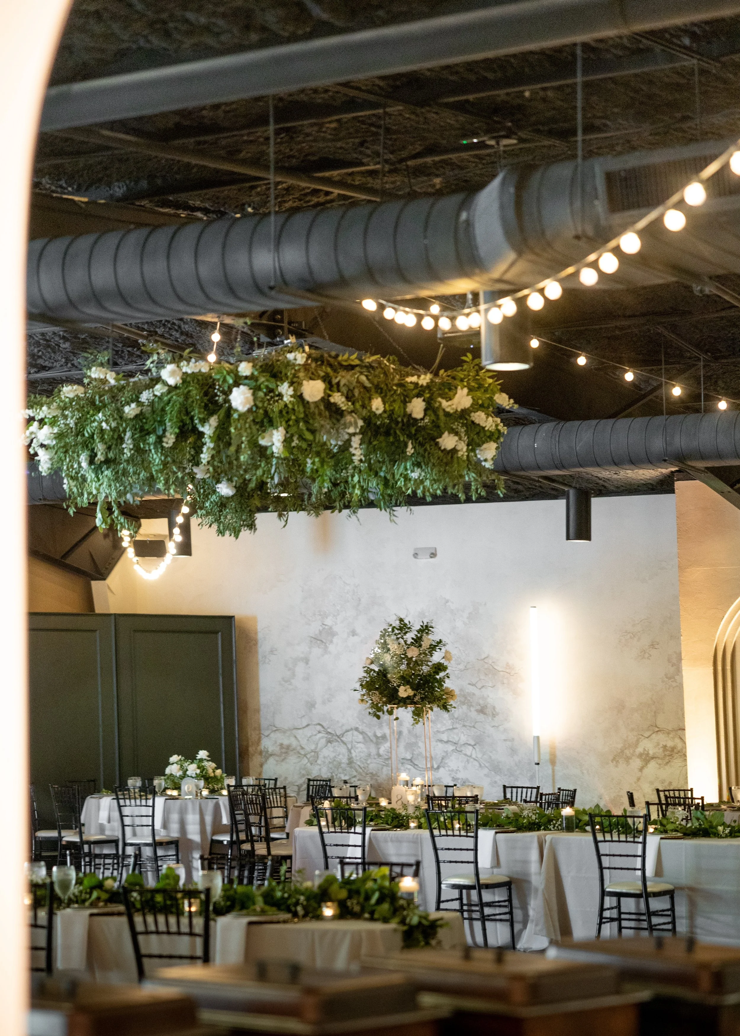 A decorated banquet hall with round tables covered in white tablecloths, surrounded by black chairs. The tables are adorned with floral centerpieces and candles. Hanging from the ceiling are string lights and large greenery arrangements with white flowers. 