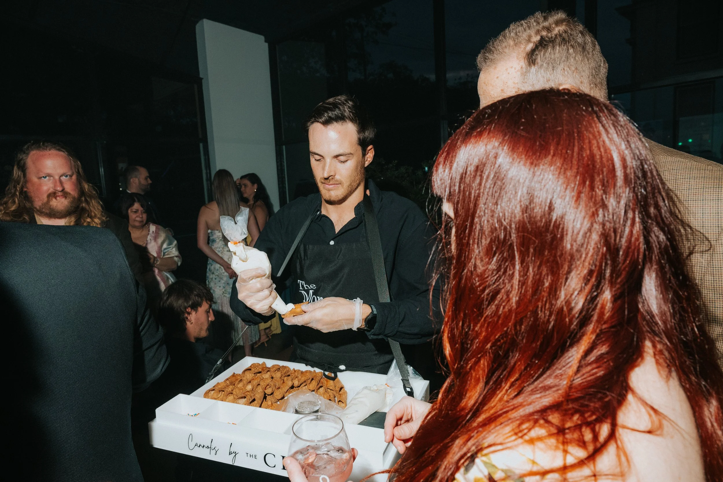 People gathered at an indoor event, with a man in a black shirt serving food from a tray of cookies or baked goods, and a woman with long red hair holding a glass of water, in front of a table with a sign that partially reads "Cannoli by the Clyde".