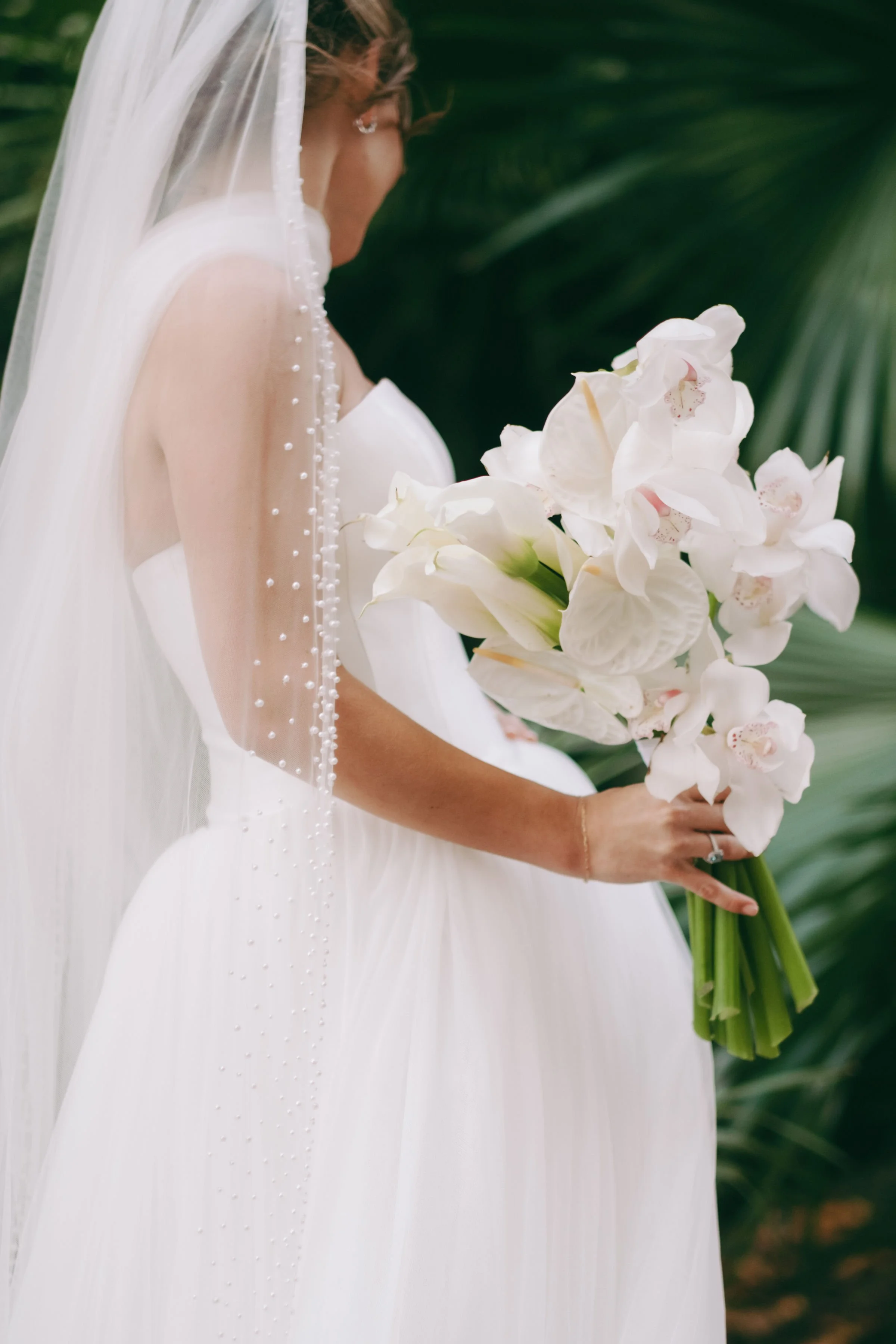 A bride in a white wedding dress holding a bouquet of white orchids. She is wearing a veil and earrings, standing against a background of green palm leaves.