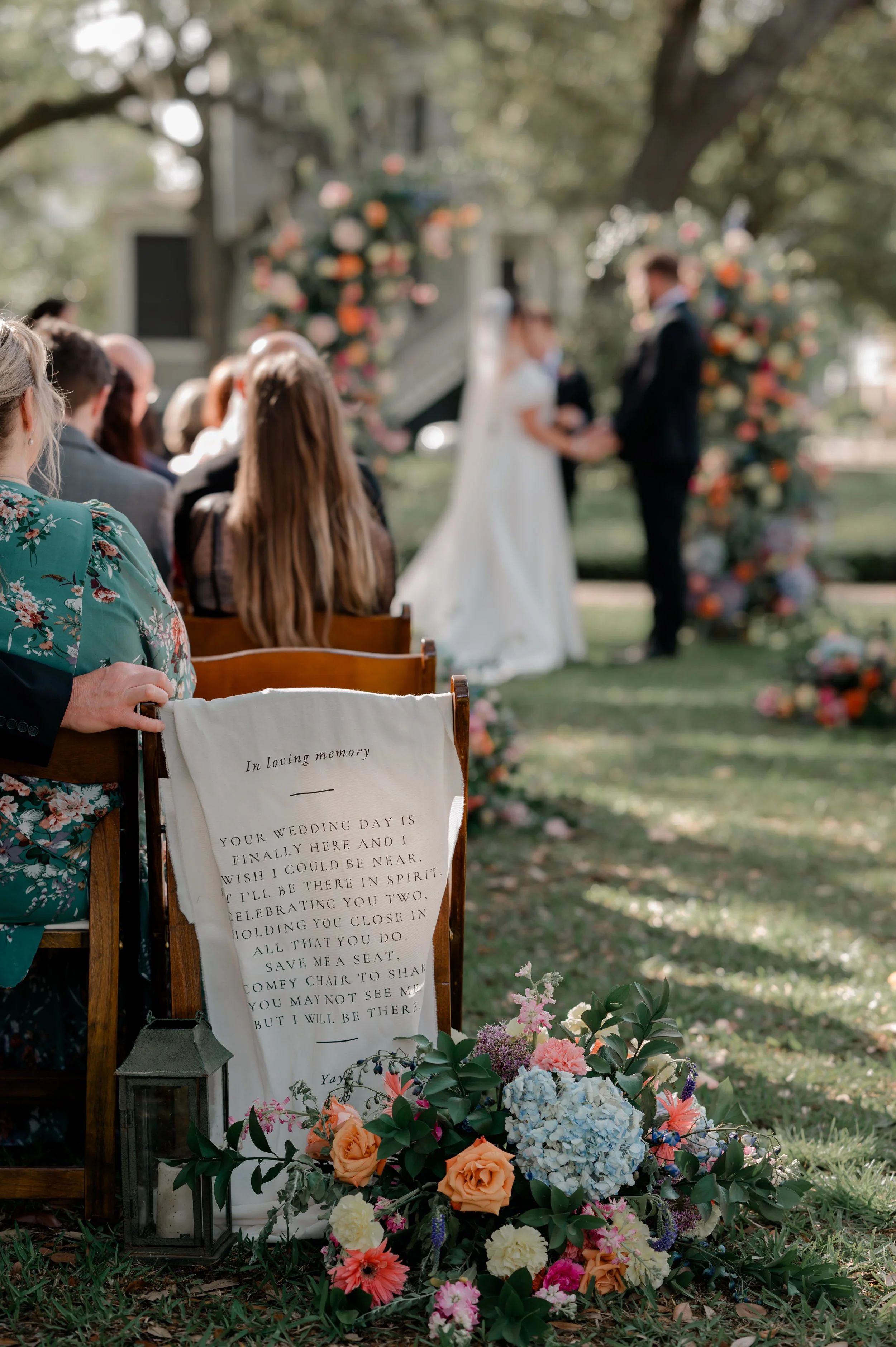 A wedding ceremony outdoors with a bride and groom holding hands, standing in front of a floral arch. Guests are seated nearby, and there are flowers, lanterns, and greenery surrounding the area.