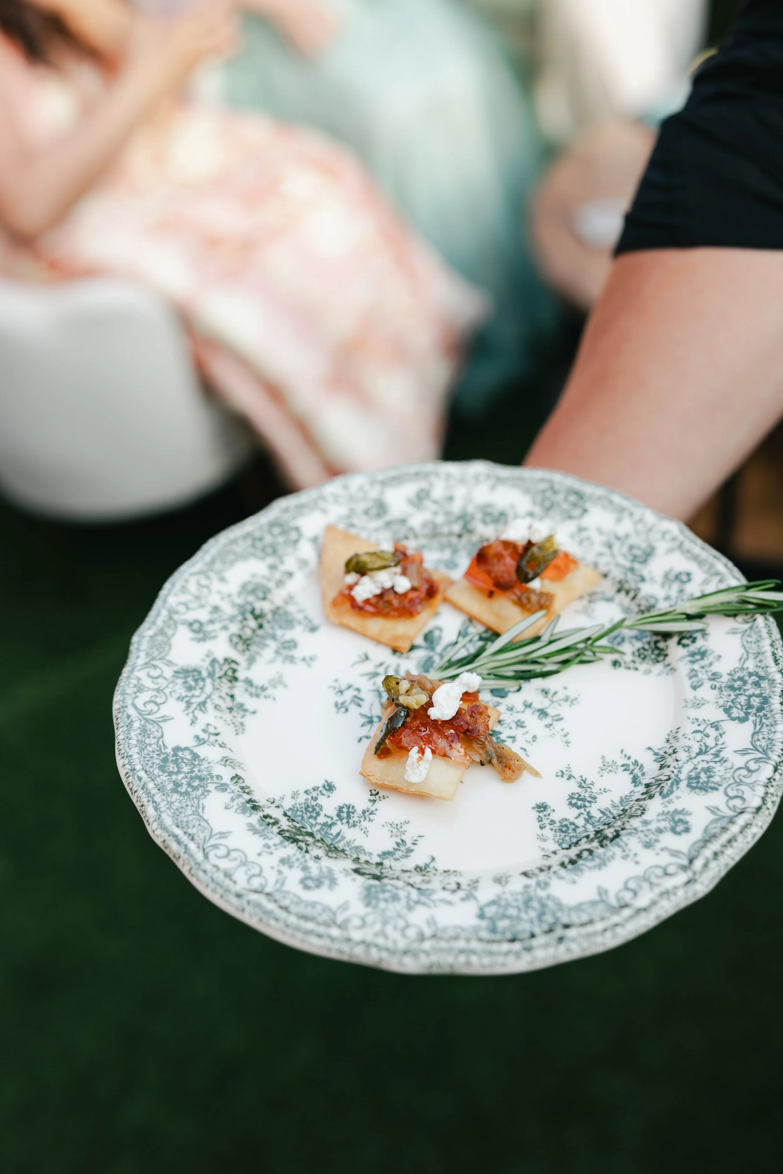A close-up of a decorative plate with four small, rectangular food appetizers topped with vegetables and cheese, held by a person in black clothing. The background is blurred, showing a person in floral attire and green outdoor setting.