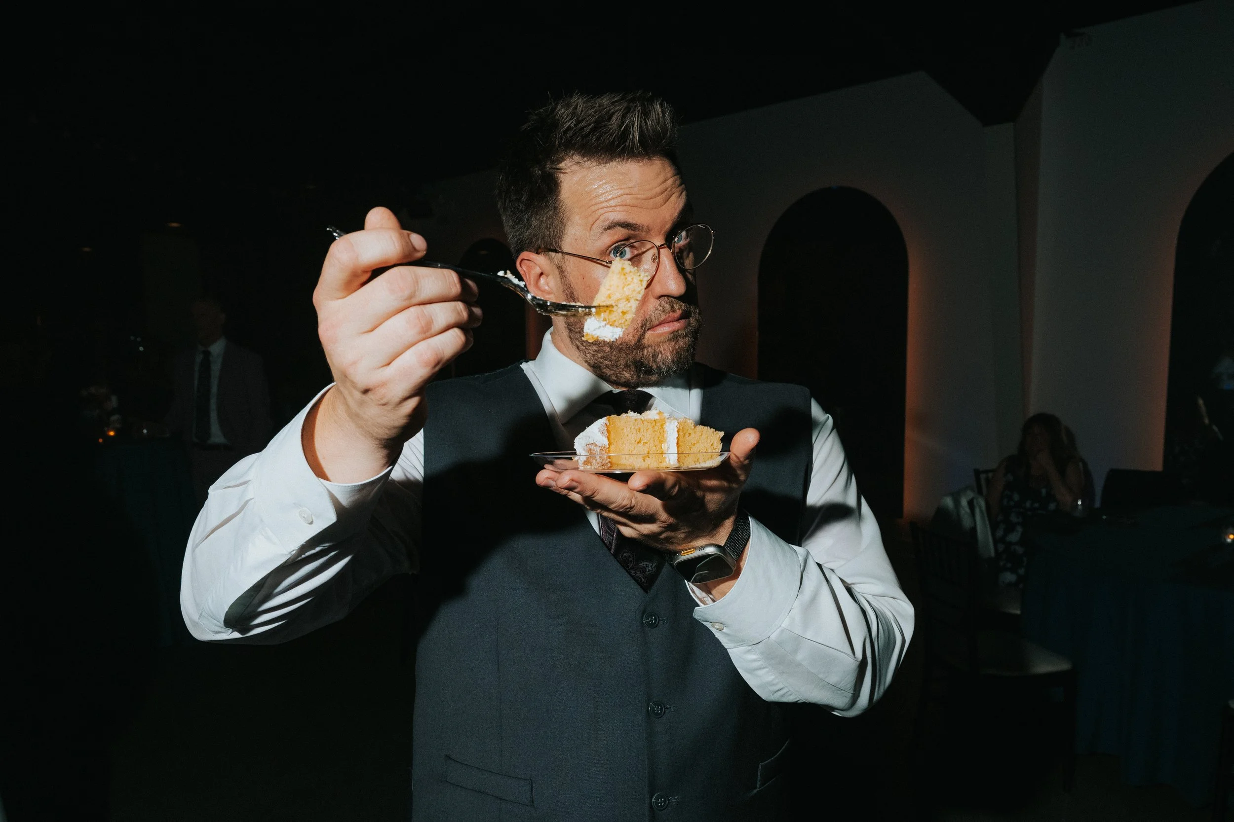 A man dressed in formal attire, wearing glasses, is holding a fork with a piece of cake and a small plate with more cake in a dimly lit setting, possibly at a wedding or event.