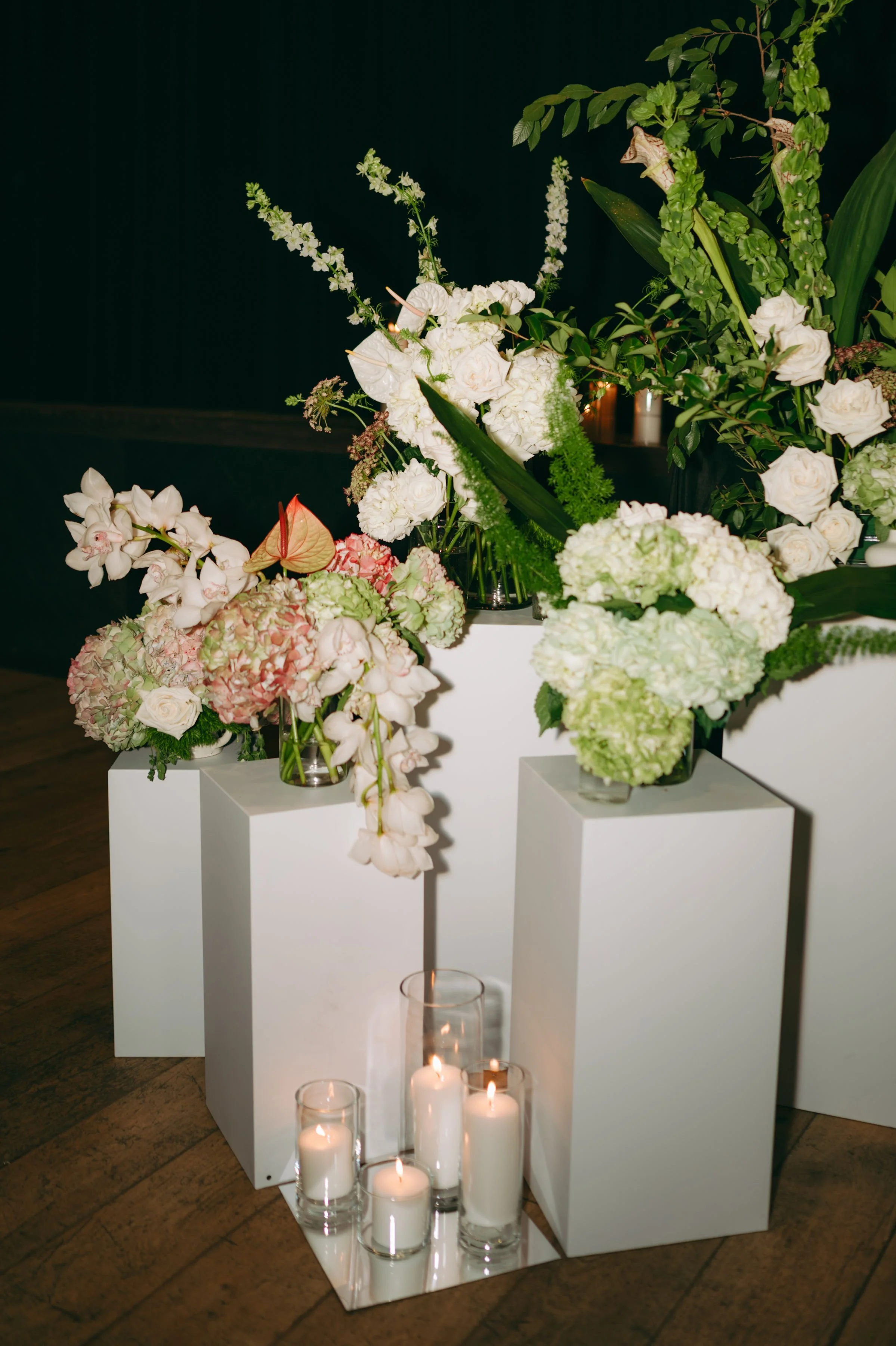 Elegant floral arrangement featuring white roses, hydrangeas, orchids, and greenery on a white display stand, with candles in glass holders at the base, against a dark background.