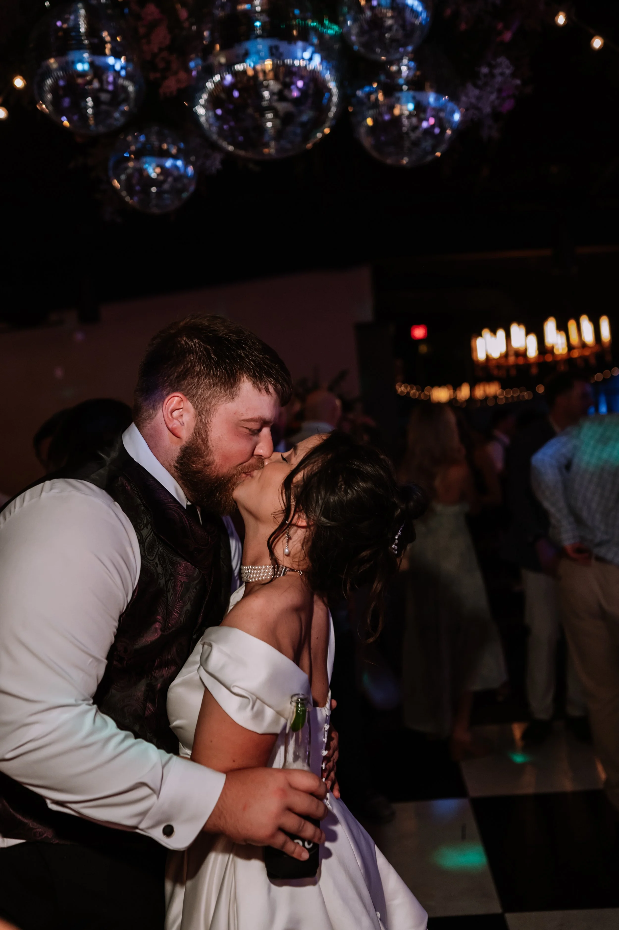 A couple is kissing at a celebration event, with the man wearing a white dress shirt and a dark vest, and the woman in a white dress with puffy sleeves, wearing pearl jewelry, holding a drink. The background shows other people and hanging disco balls