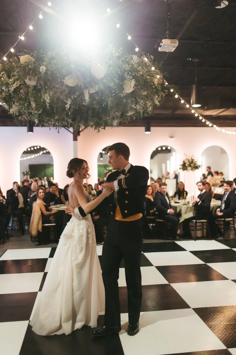 A bride and groom dancing at their wedding reception with guests seated at tables in the background, under a ceiling decorated with hanging lights and a floral arrangement.