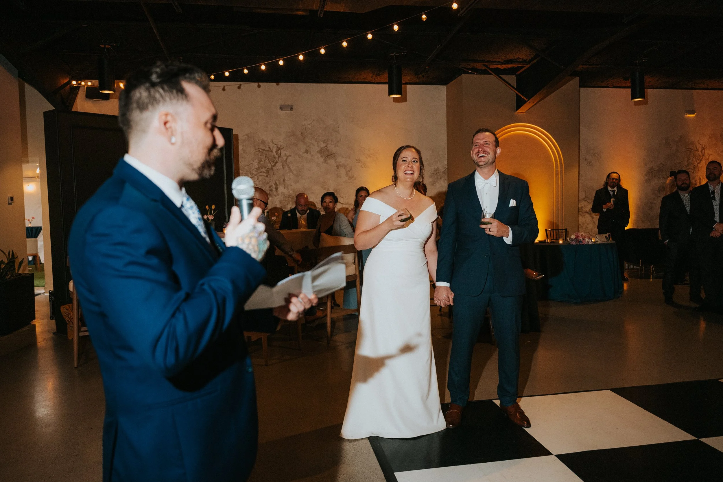 A wedding reception with a bride and groom holding hands, smiling, and listening to a man in a blue suit giving a speech or toast. Guests are seated at tables in the background, and the room is decorated with warm lighting and string lights.