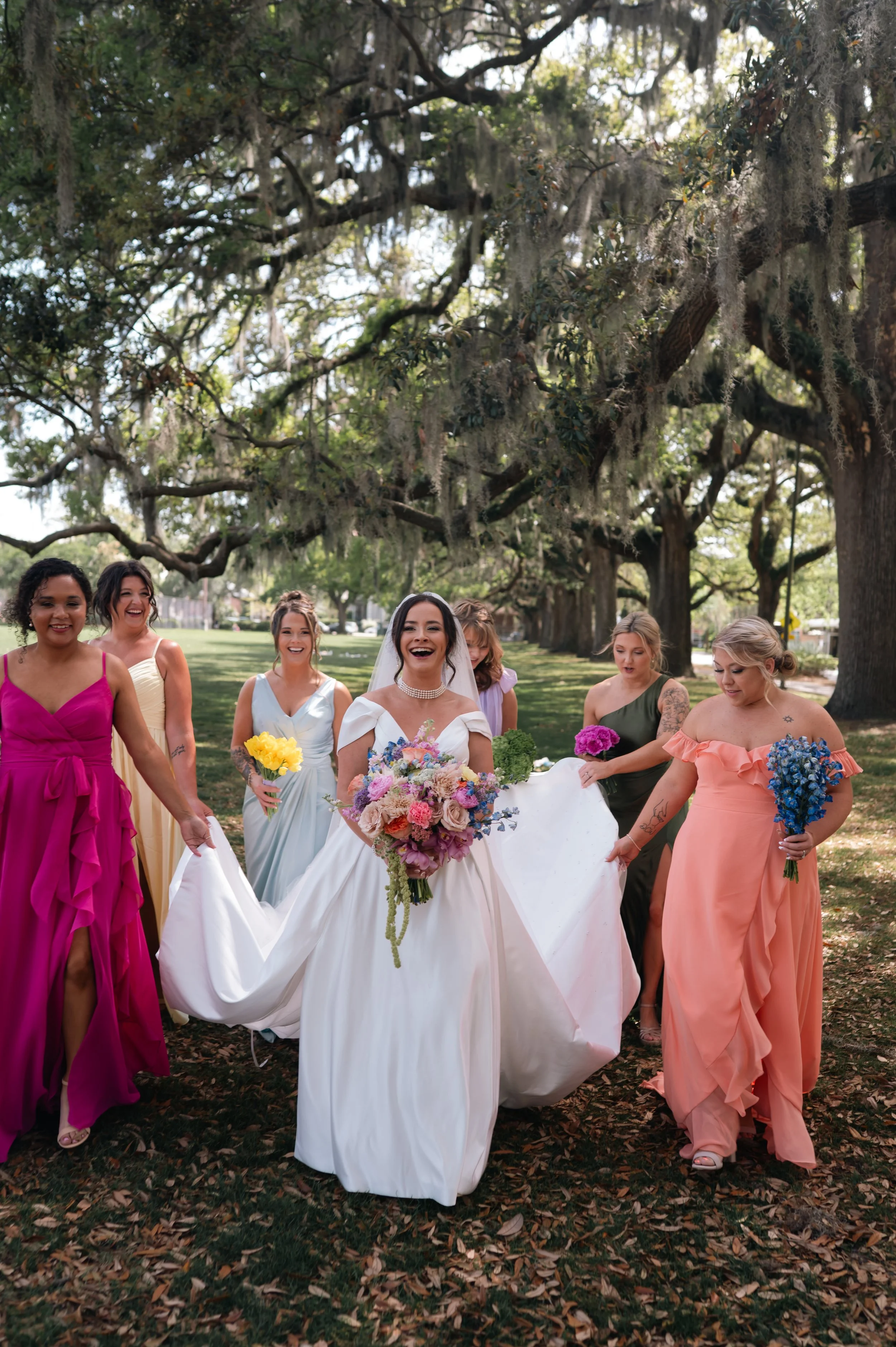 A group of women, including a bride in a white wedding dress holding a bouquet of flowers, walking outdoors under large trees with Spanish moss in a park or garden setting.