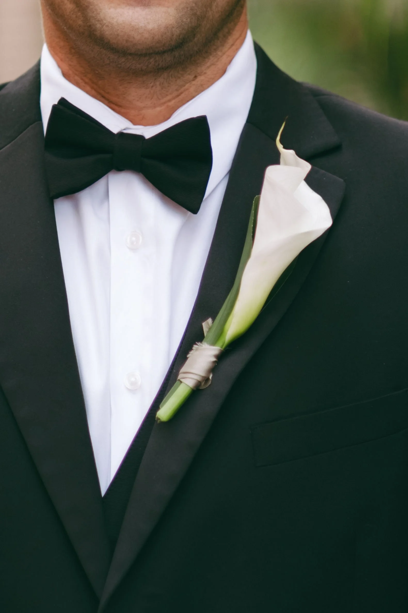 Close-up of a man wearing a black tuxedo with a white dress shirt, black bow tie, and a white calla lily boutonniere on his left lapel.