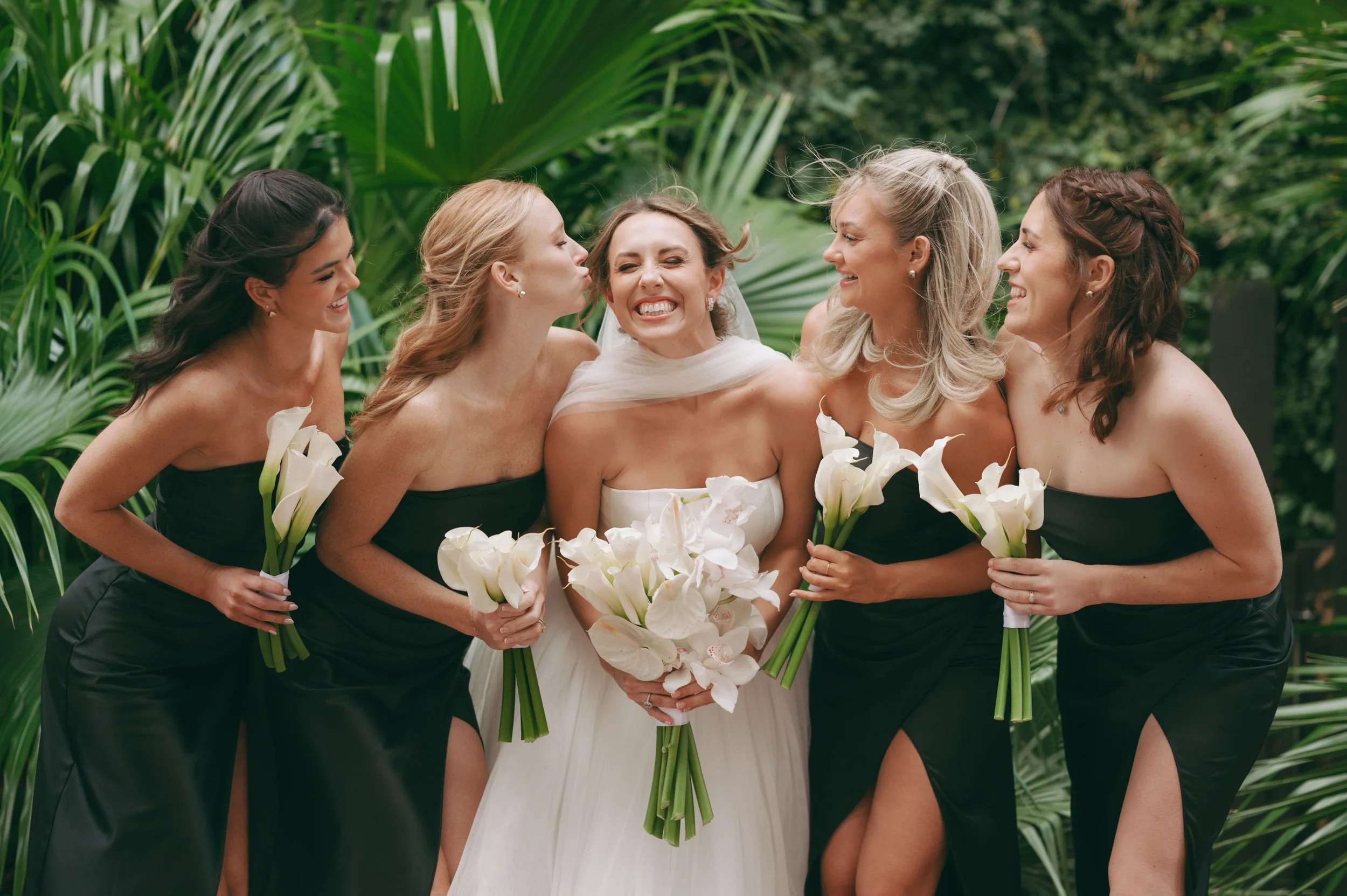 Group of women in wedding attire holding calla lily bouquets, celebrating outdoors with lush green foliage in the background.