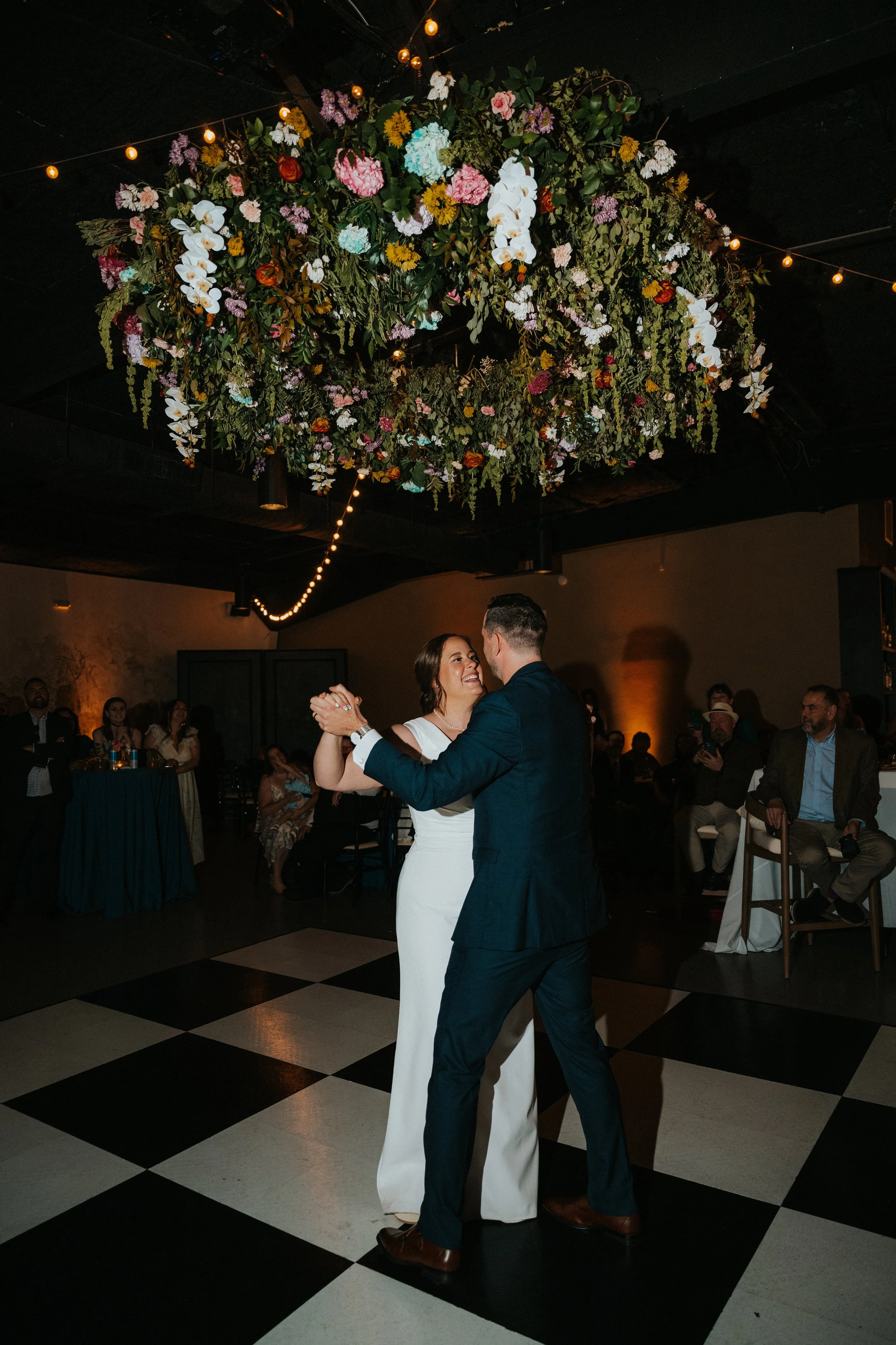 A couple dancing at a wedding reception, with the bride in a white dress and the groom in a dark suit, under a large floral arrangement with string lights overhead, and guests seated in the background.
