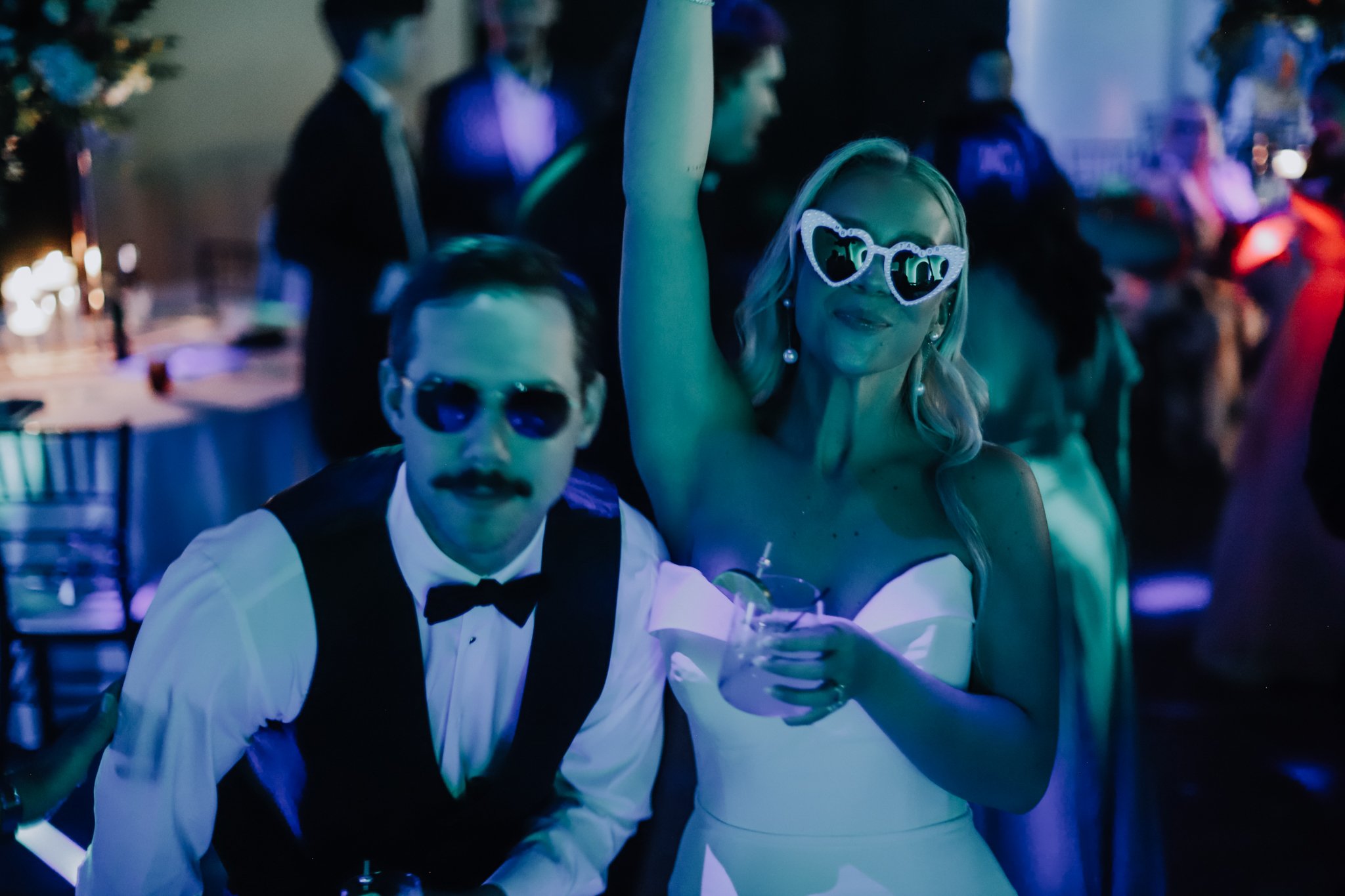 A woman wearing sunglasses with heart-shaped frames and pearl earrings dancing at a party. A man dressed as a waiter with sunglasses, a white shirt, and a black vest is sitting beside her, holding a drink with a straw in his hand. The background show