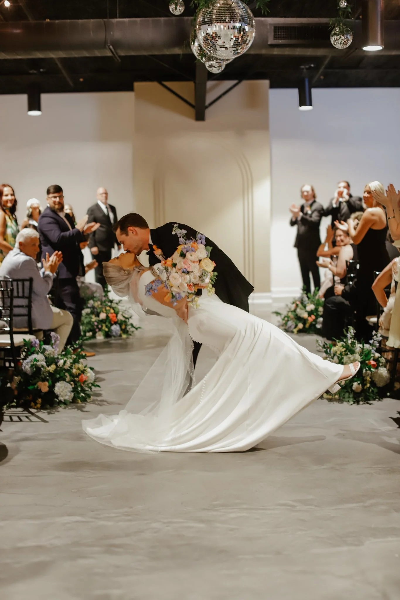 A newlywed couple sharing a romantic dance at their wedding reception, surrounded by guests clapping and smiling, with floral arrangements and a disco ball hanging from the ceiling.