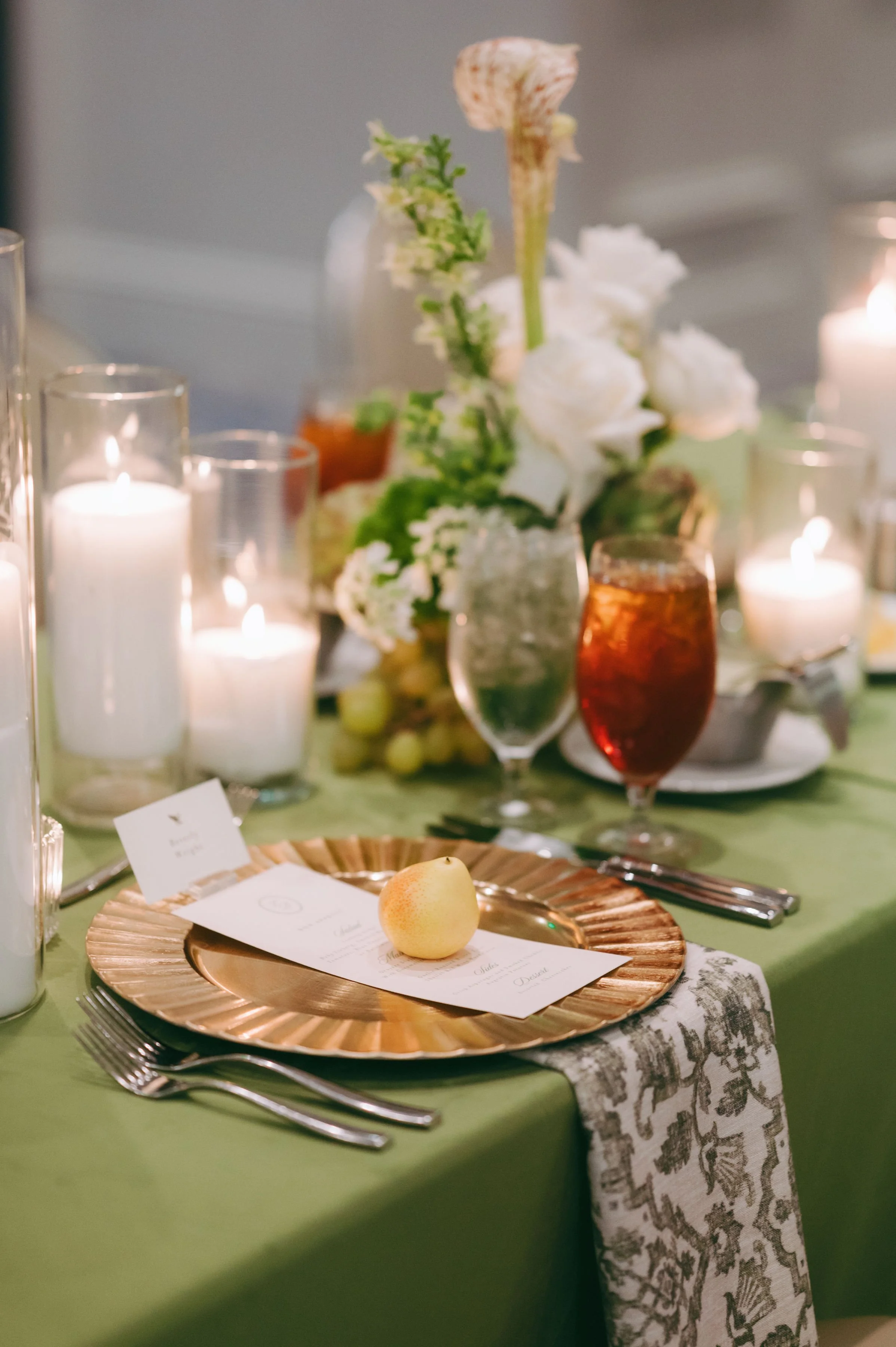 A table setting with a gold decorative charger plate, a peeled pear on top, and a menu card. The table has a green tablecloth and a patterned table runner. In the background, there are white candles and floral arrangements.
