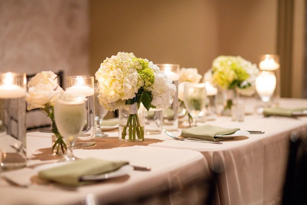 Elegant dining table with white tablecloth, floral centerpieces, candles, and place settings in a softly lit room.
