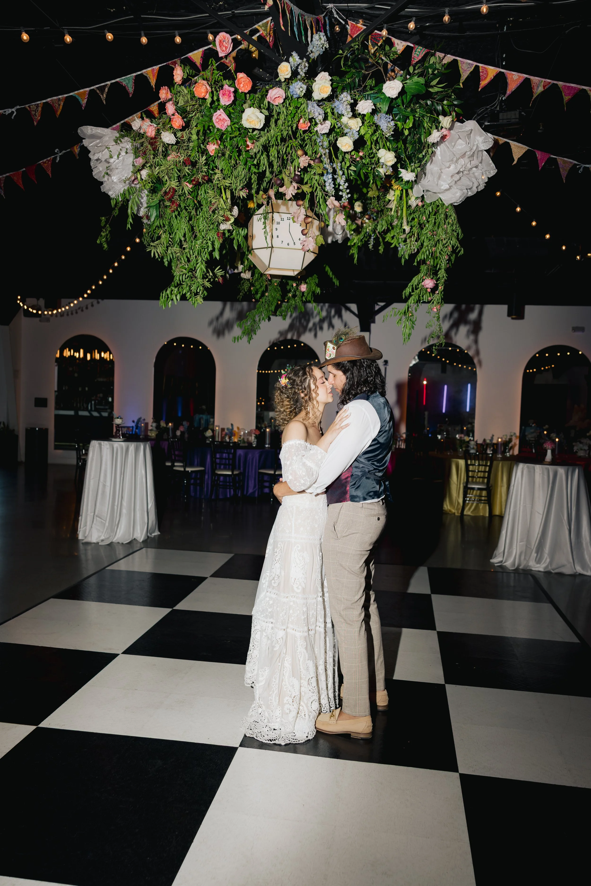 A bride and groom share a dance at their wedding reception in an indoor venue with black and white checkered flooring and a large floral decoration hanging from the ceiling. The bride wears a white lace wedding dress and has curly hair, while the gro
