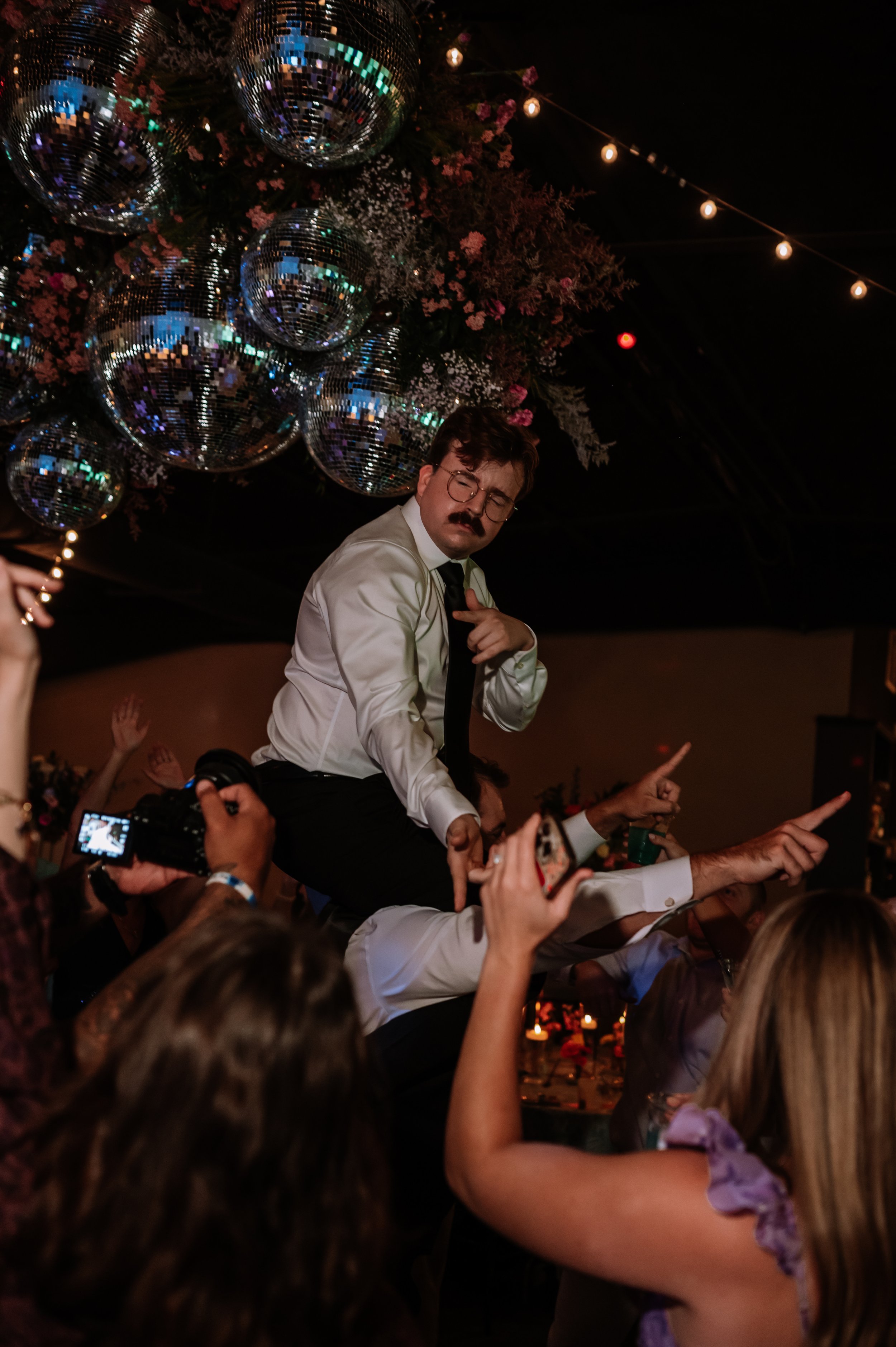 A man with glasses and a mustache is dancing on the shoulders of another person in a crowded party with disco balls, pink flowers, and string lights.
