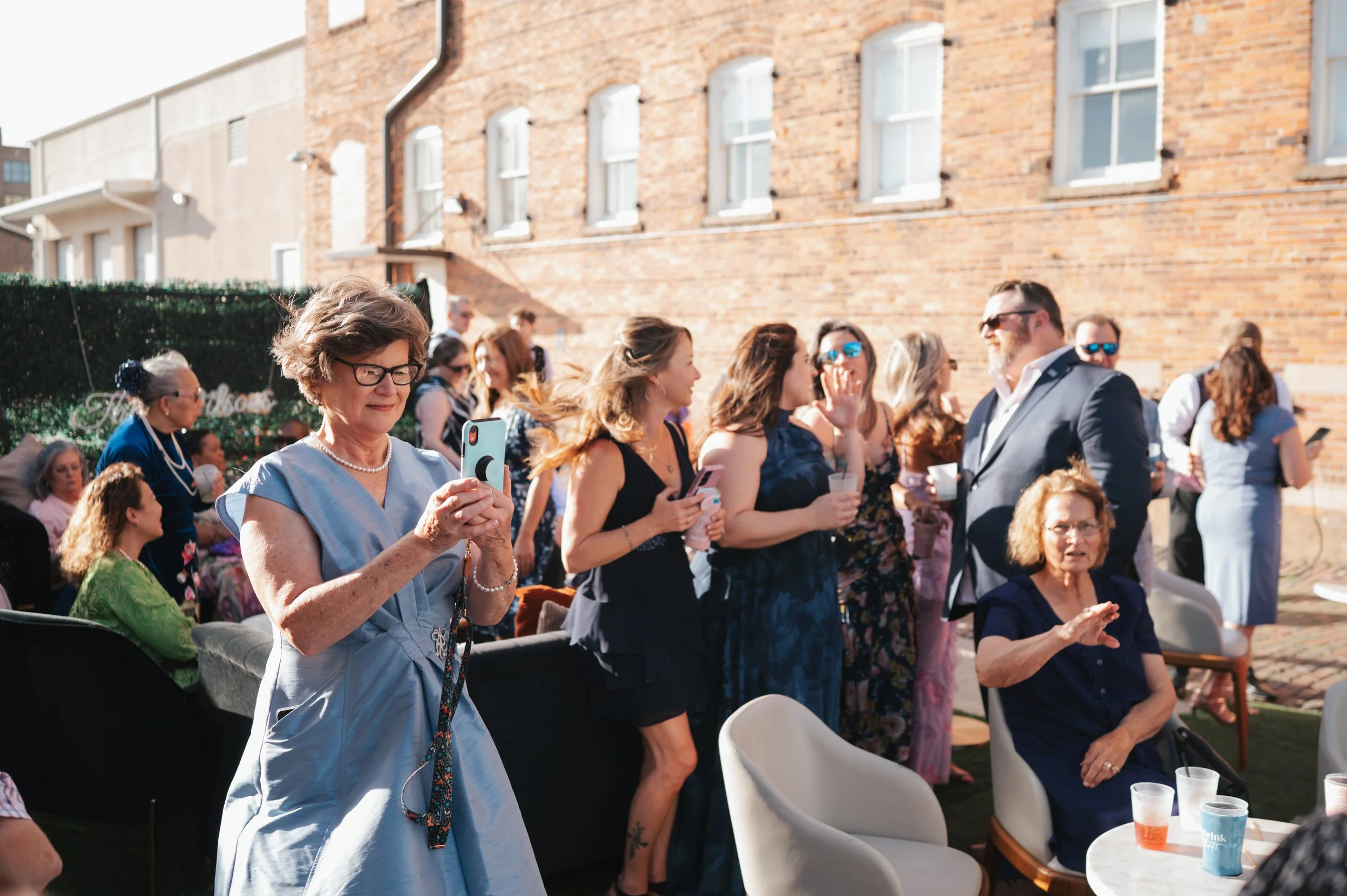 A woman with glasses and a pearl necklace wearing a light blue dress is capturing a photo of the cocktail hour. There are multiple women and a man in formal attire interacting and socializing outdoors against a brick building during daytime.