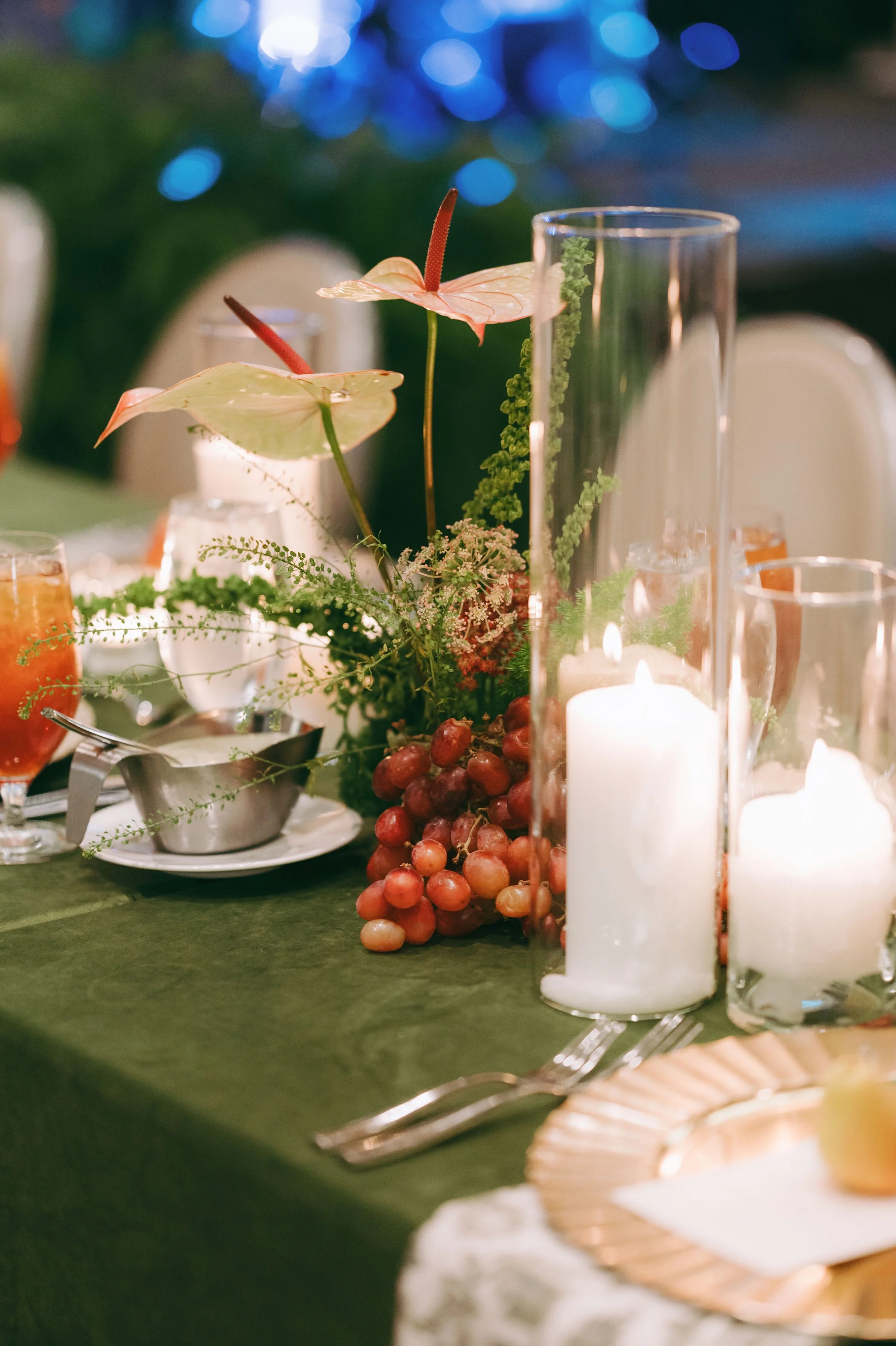 Close-up of a table centerpiece featuring pink anthurium flowers, green foliage, red grapes, and white candles in glass holders at a dinner event.