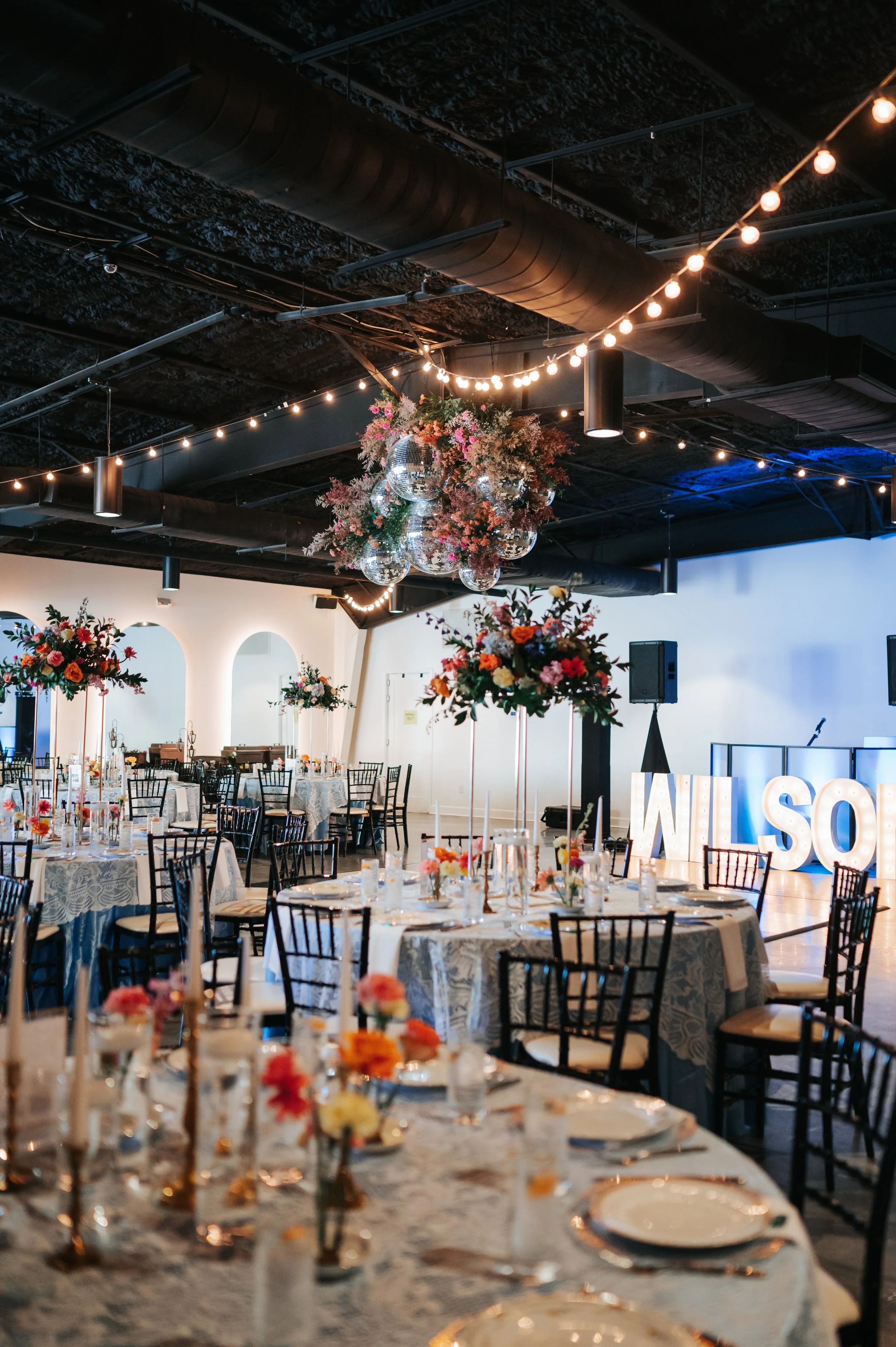 Interior of a decorated event venue with floral centerpieces on tables, string lights hanging from the ceiling, and disco balls as a centerpiece of a floral arrangement. There is a large customized illuminated sign in the background.
