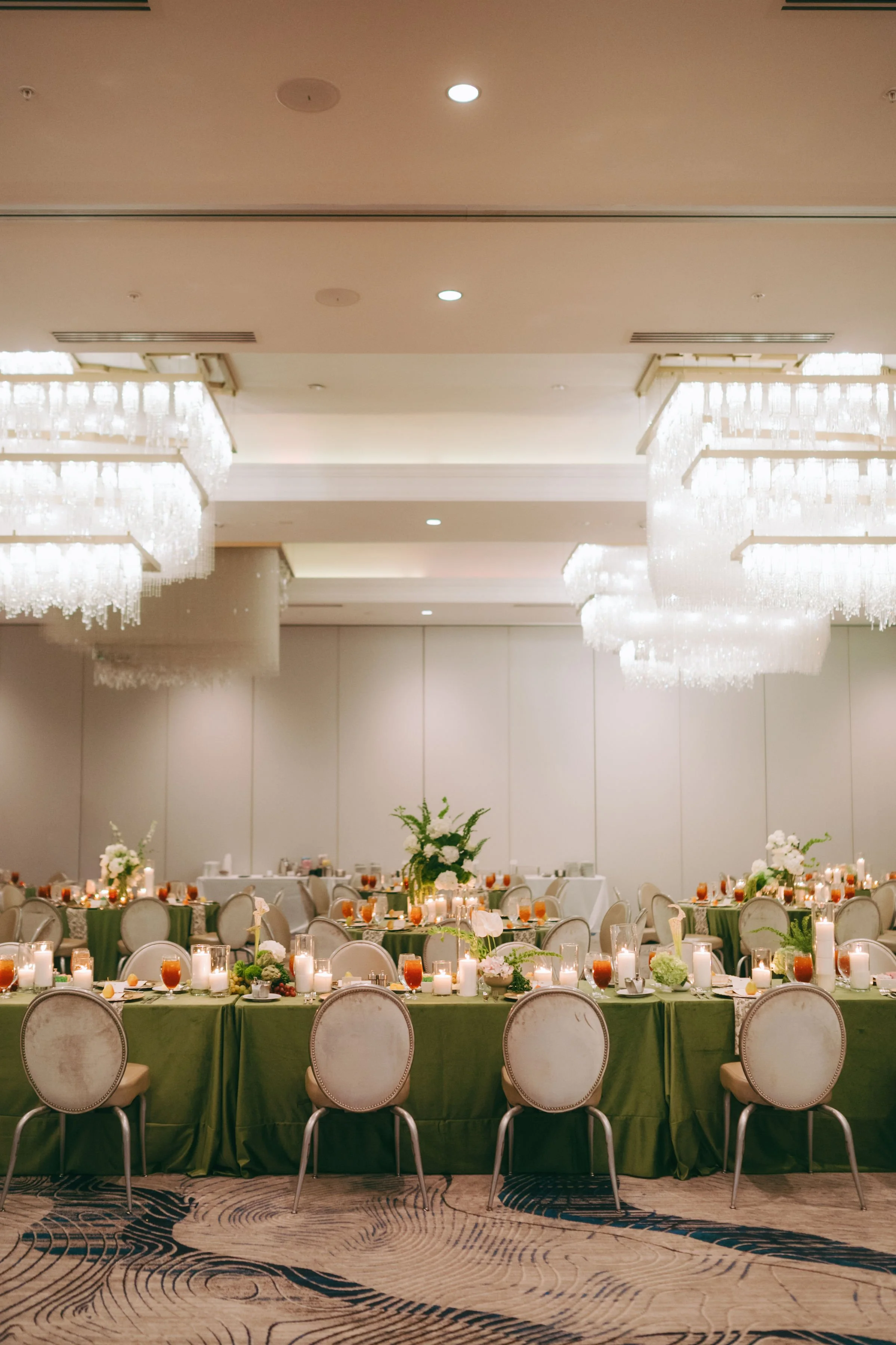 A banquet hall decorated with elegant chandeliers, round tables with green tablecloths, and floral centerpieces. The top portion of the image shows a ceiling with recessed lighting, while the lower part shows a patterned carpet and chairs around the 