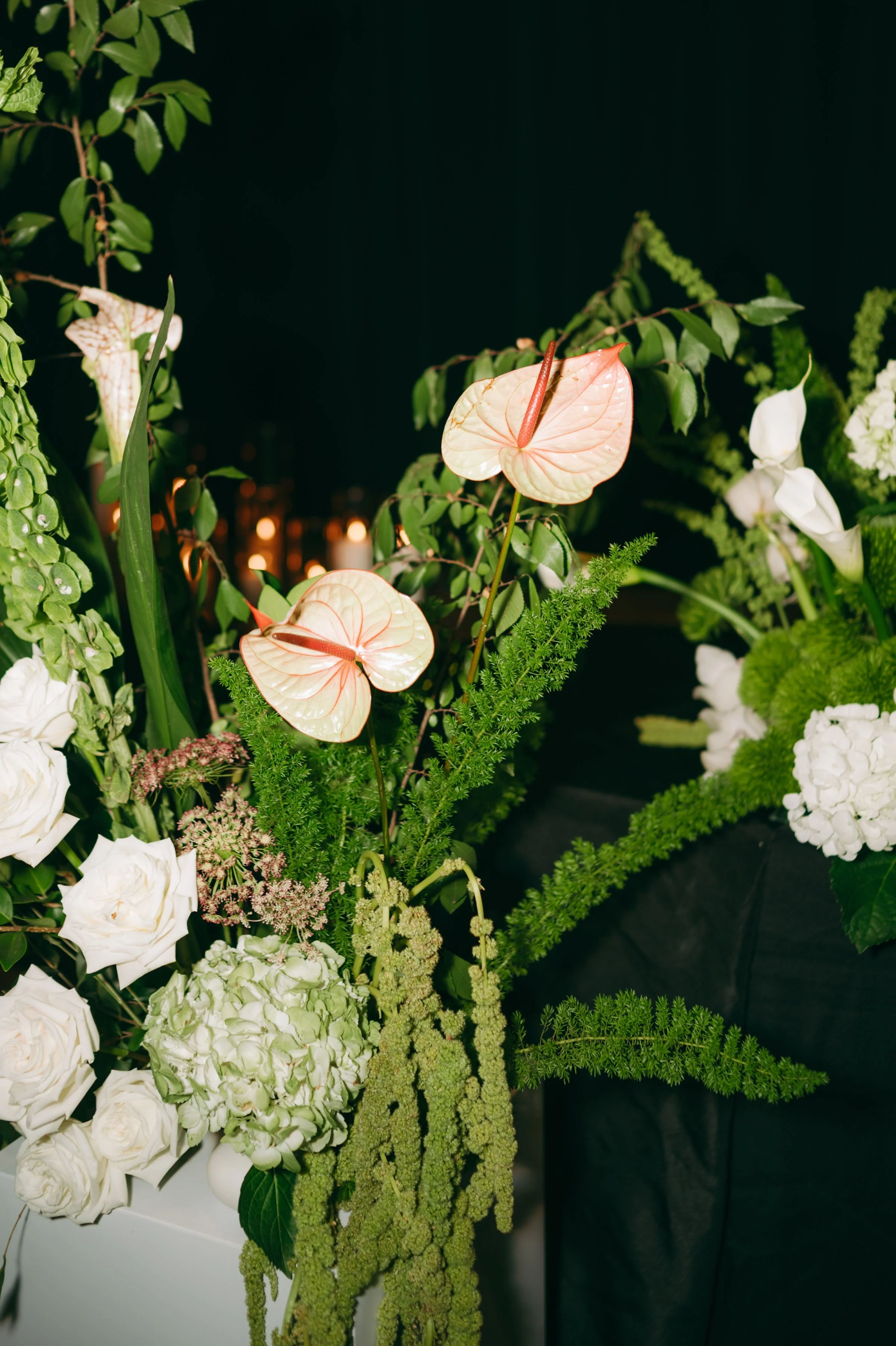 Close-up of a floral arrangement with white roses, white hydrangeas, green foliage, and anthurium flowers with pinkish spathes and greenish spadixes, set against a dark background.