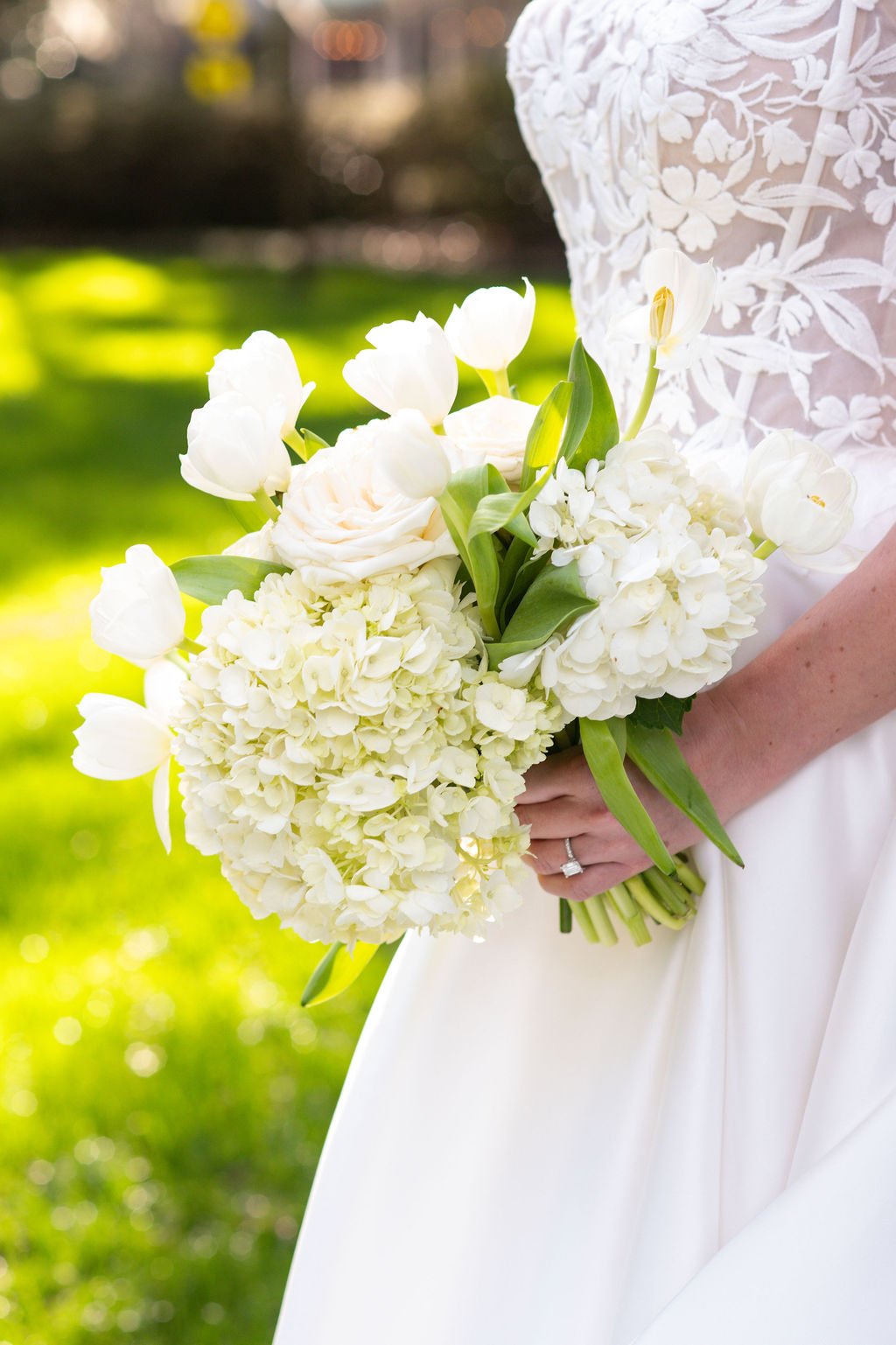 A bride in a white dress holding a bouquet of white flowers, including tulips, roses, hydrangeas, and other blooms, outdoors with a blurred green background.