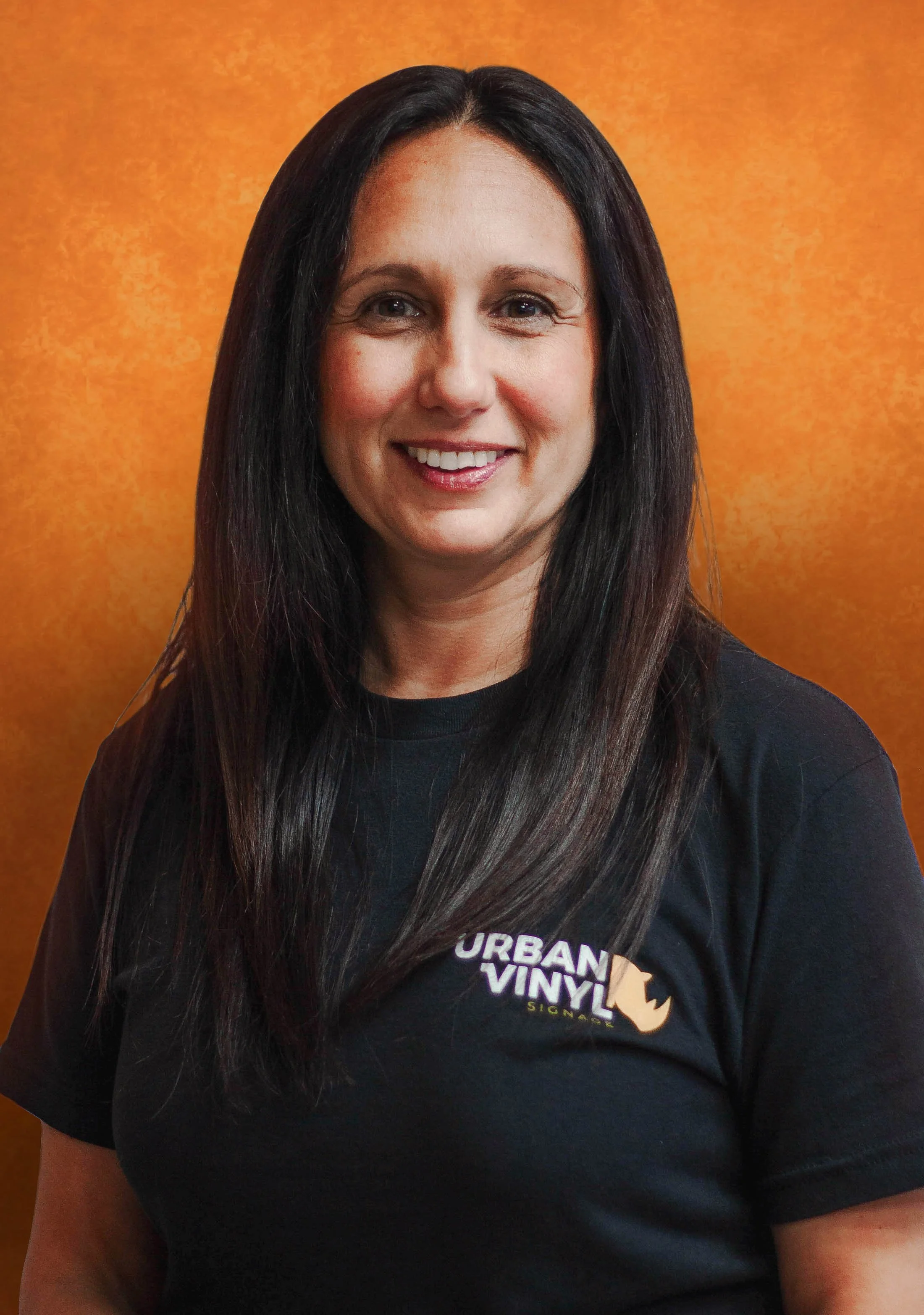 Woman with glasses smiling while standing outdoors, wearing a black top and dark hair, with a blurred building in the background.