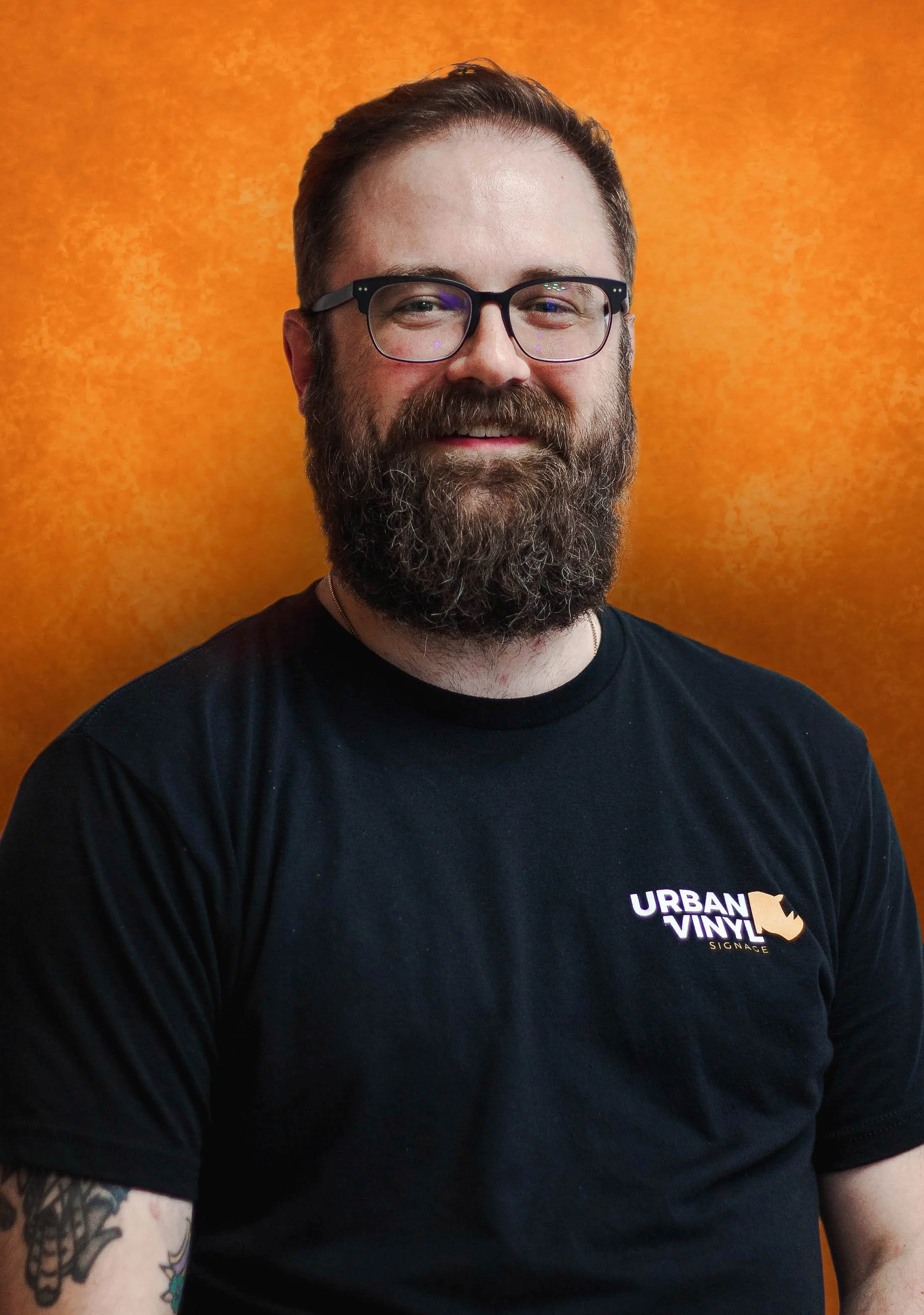Man with beard and glasses wearing an Urban Vinyl Signage sweatshirt, standing outside a building.