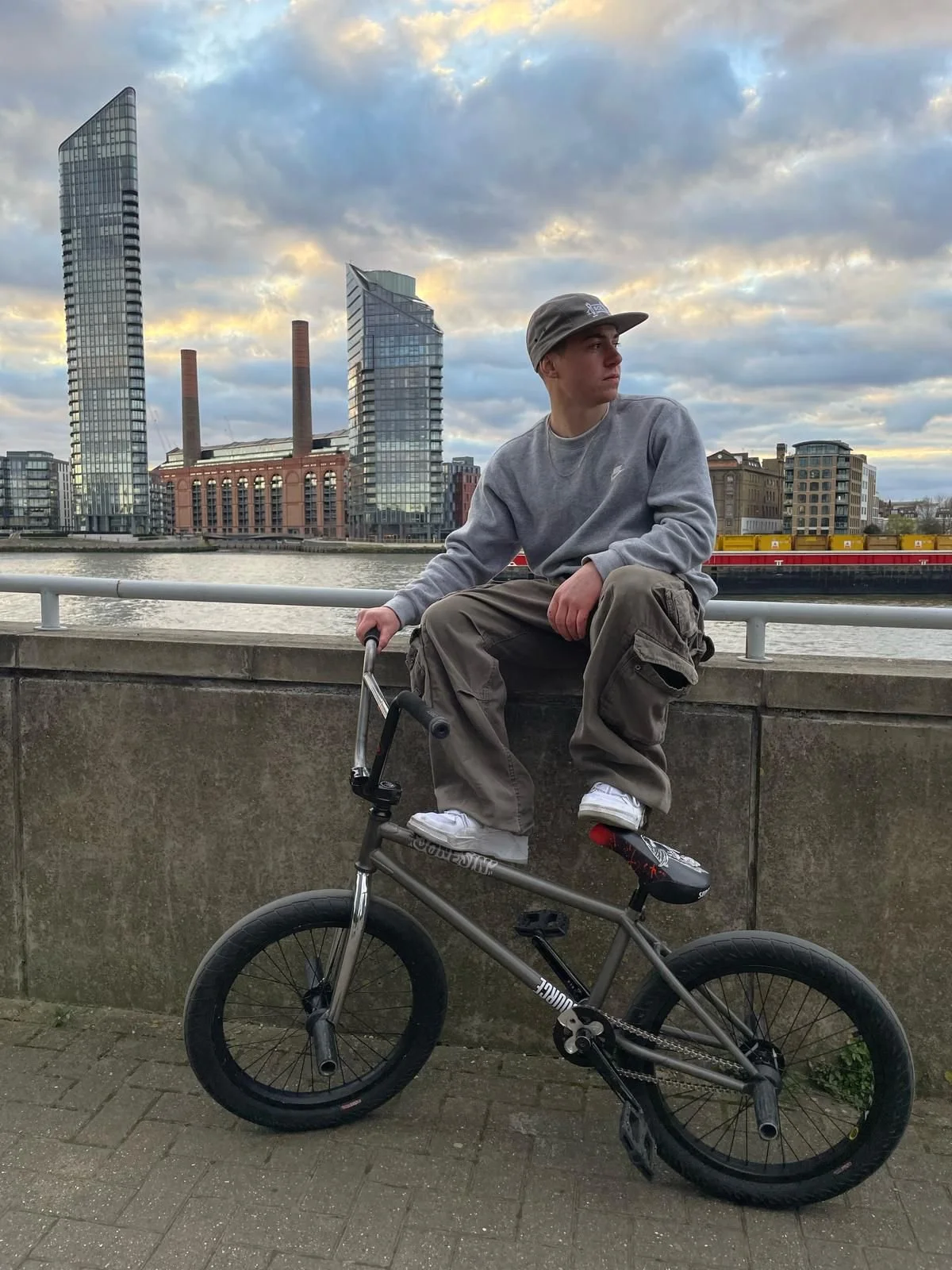 A person sitting on a BMX bike in an urban setting with modern buildings in the background, under a cloudy sky.