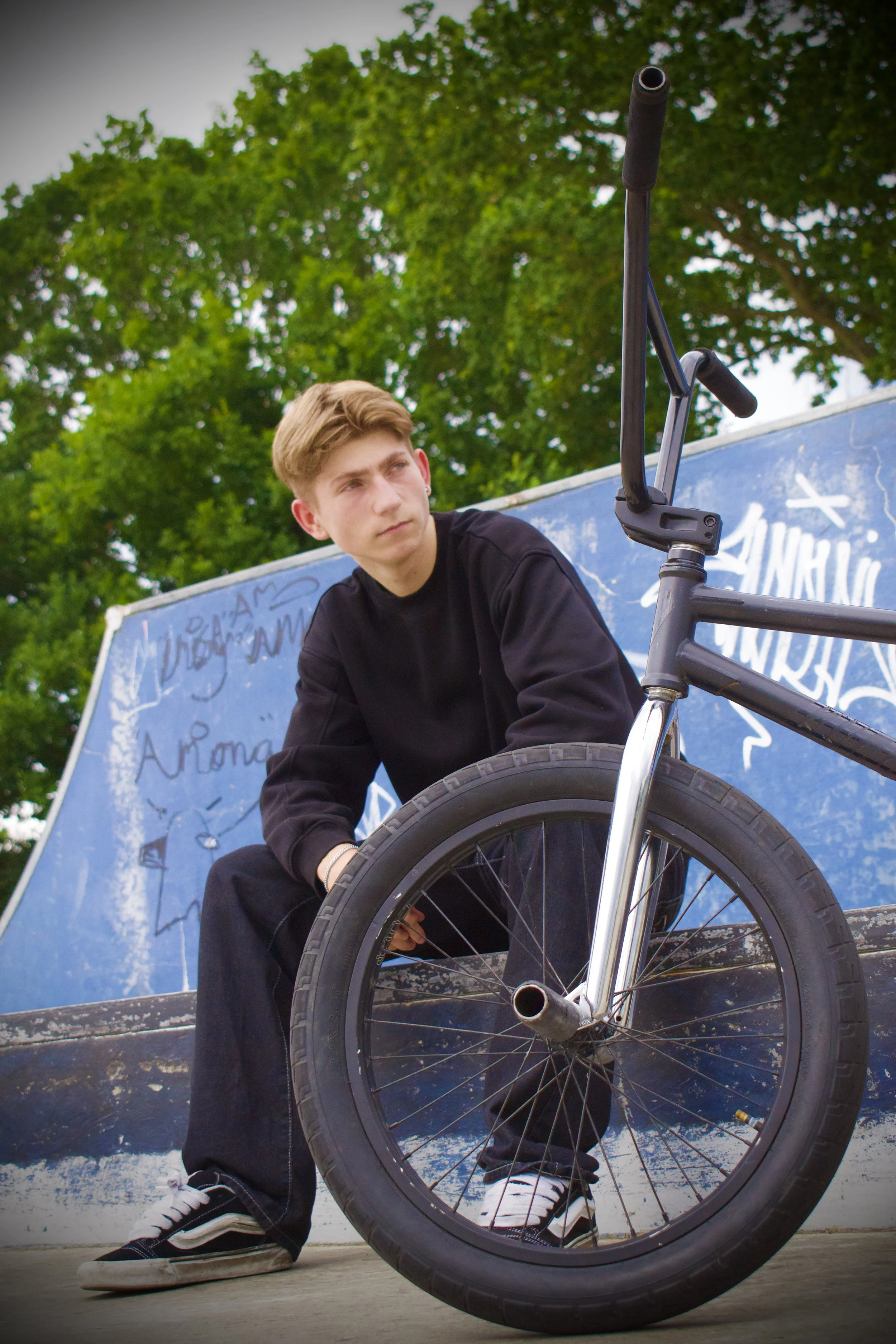 Teenager with a BMX bike at a skate park, sitting against a graffiti-covered ramp, with trees in the background.