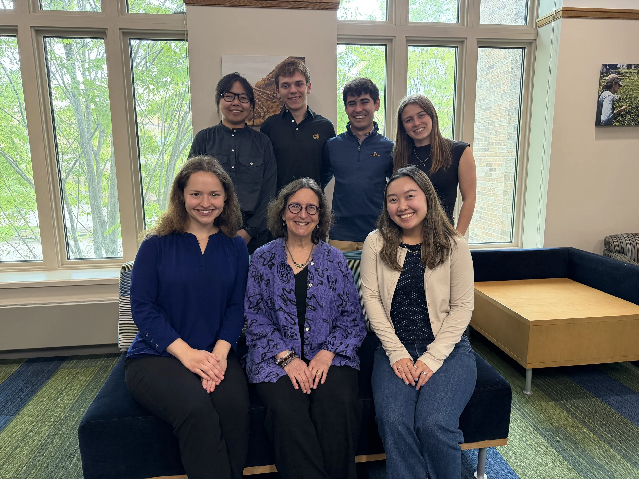 The seven members of the School Story Lab, seated and standing together in a sunlit lounge at the University of Notre Dame.