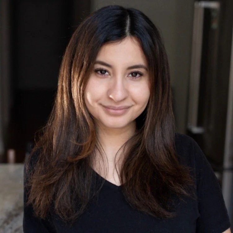 Young woman with long brown hair, smiling, wearing a black top indoors.