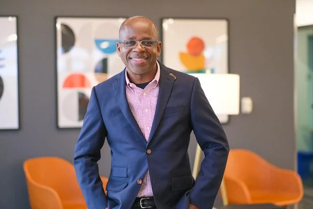 A smiling man in a navy blazer and pink checkered shirt standing in a modern office with abstract wall art and orange chairs.