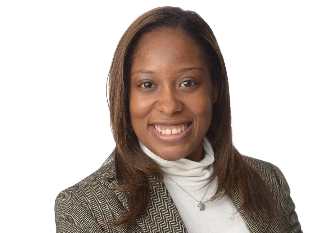 Portrait of a woman with shoulder-length brown hair, smiling, wearing a white turtleneck and a plaid blazer, against a white background.