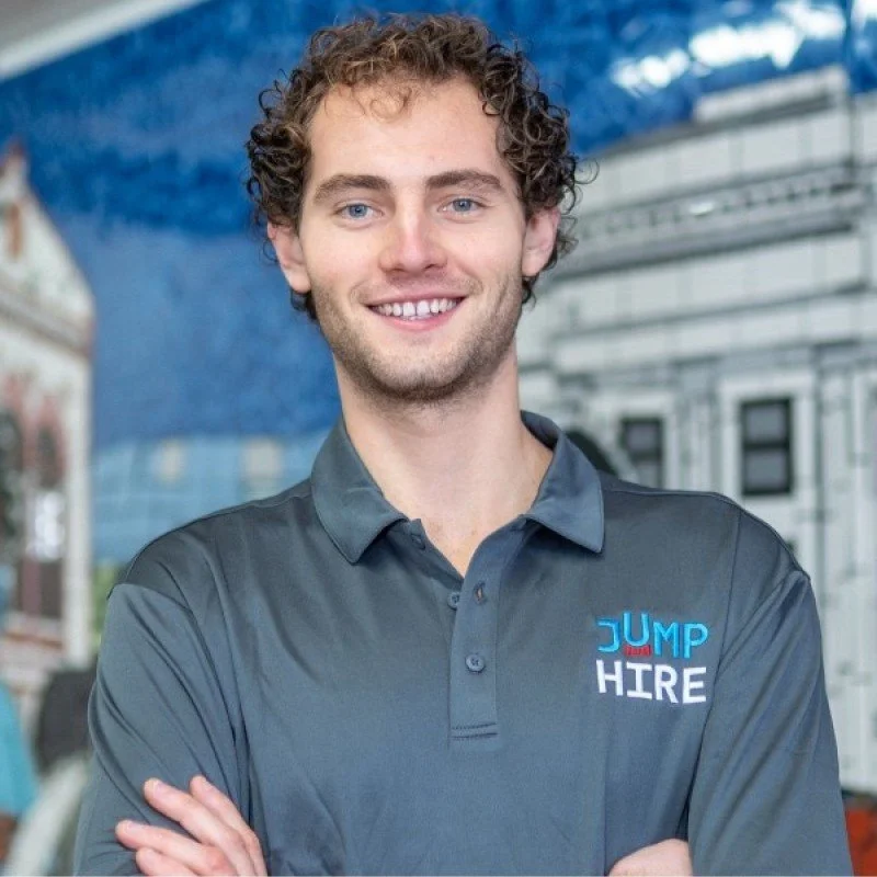 A young man with curly brown hair is smiling and standing with arms crossed in front of a backdrop of a blue sky and buildings. He is wearing a dark gray polo shirt with a logo that says 'JUMP HIRE'.
