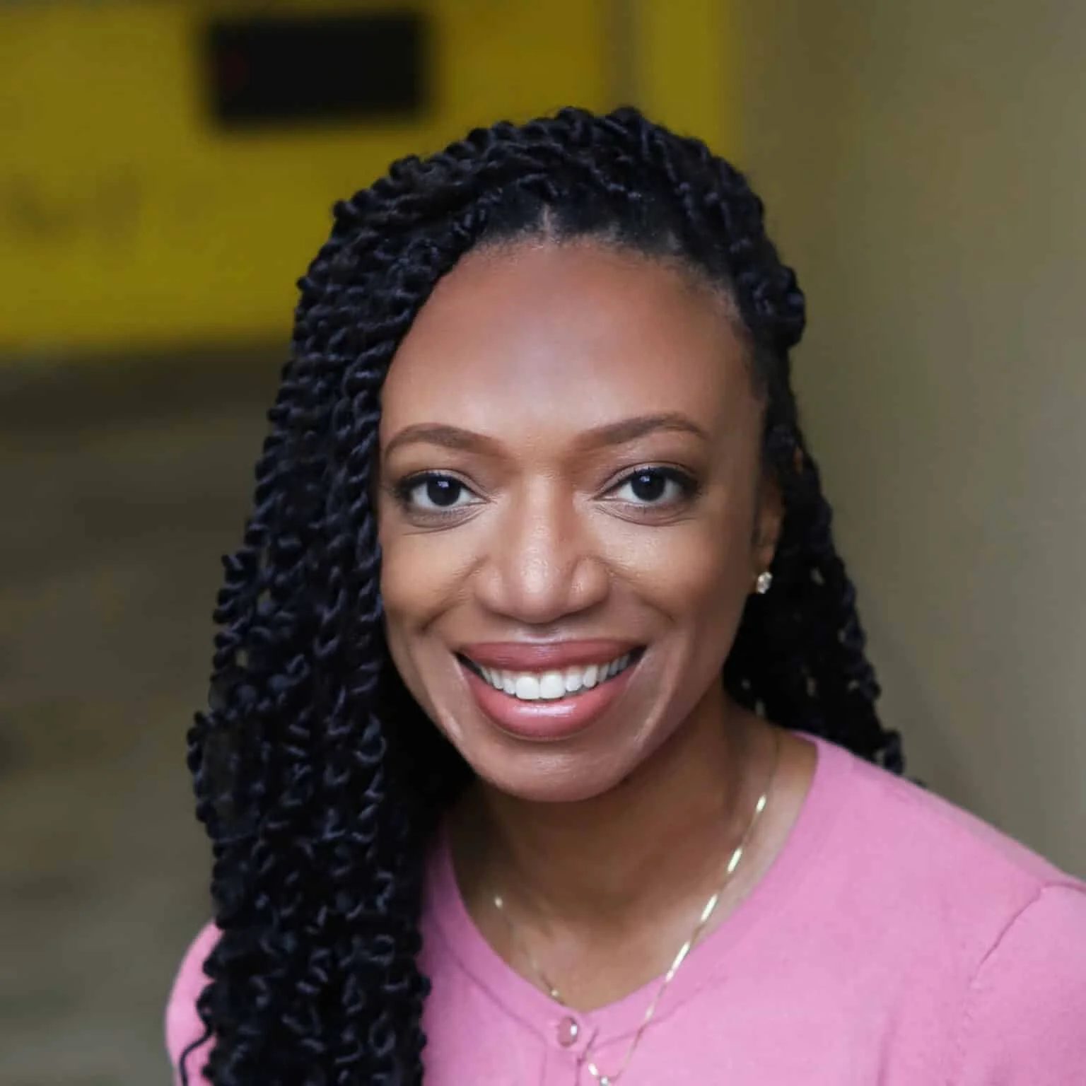 Close-up of smiling woman with braided hair, wearing pink top and jewelry, indoors.