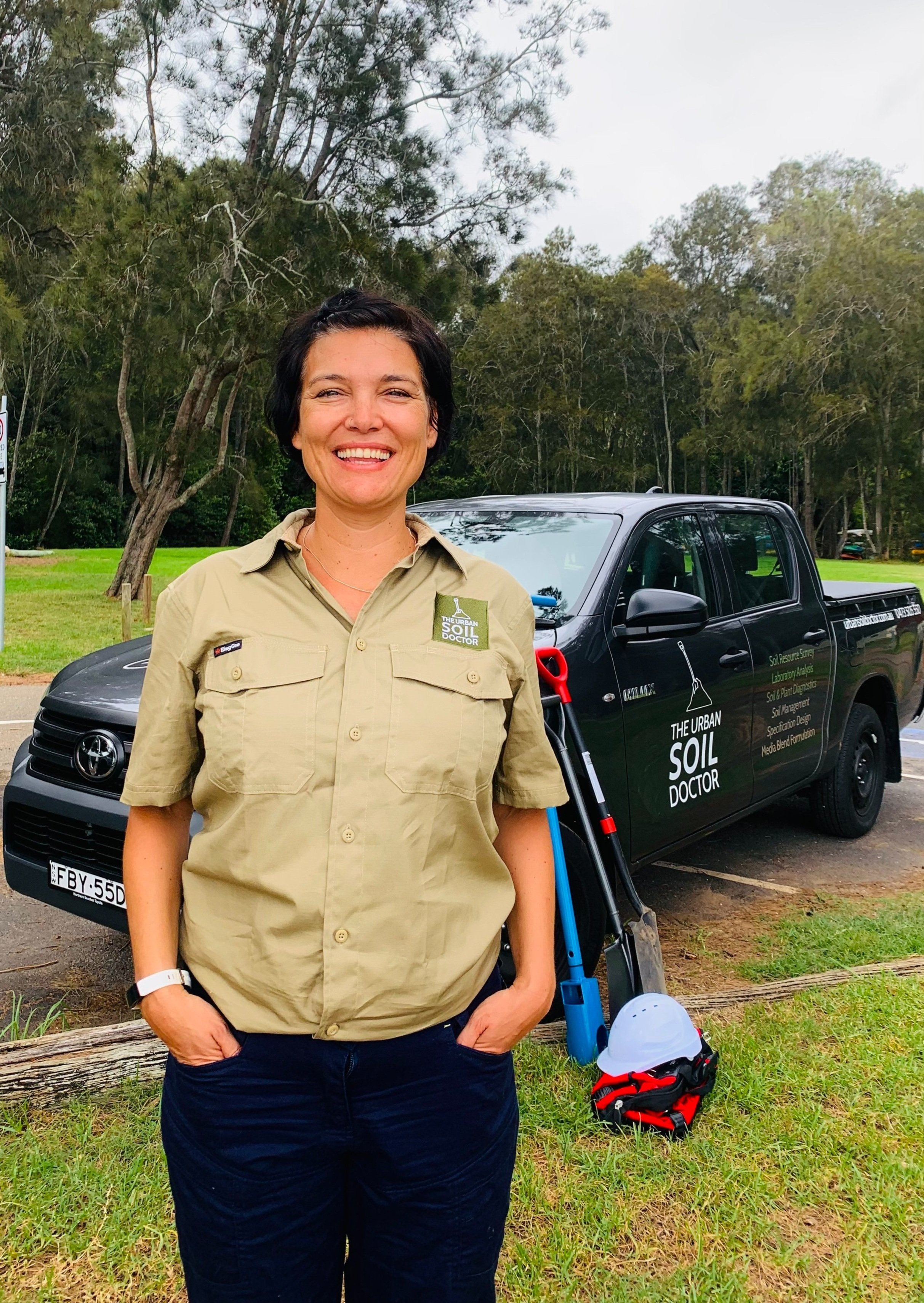 A woman smiling outdoors standing in front of a black SUV with the words "The Urban Soil Doctor" and a logo on the side. She is wearing a beige work shirt with soil-related patches, and there is a helmet, a shovel, and a pair of gloves on the ground beside her. The background features trees and an overcast sky.
