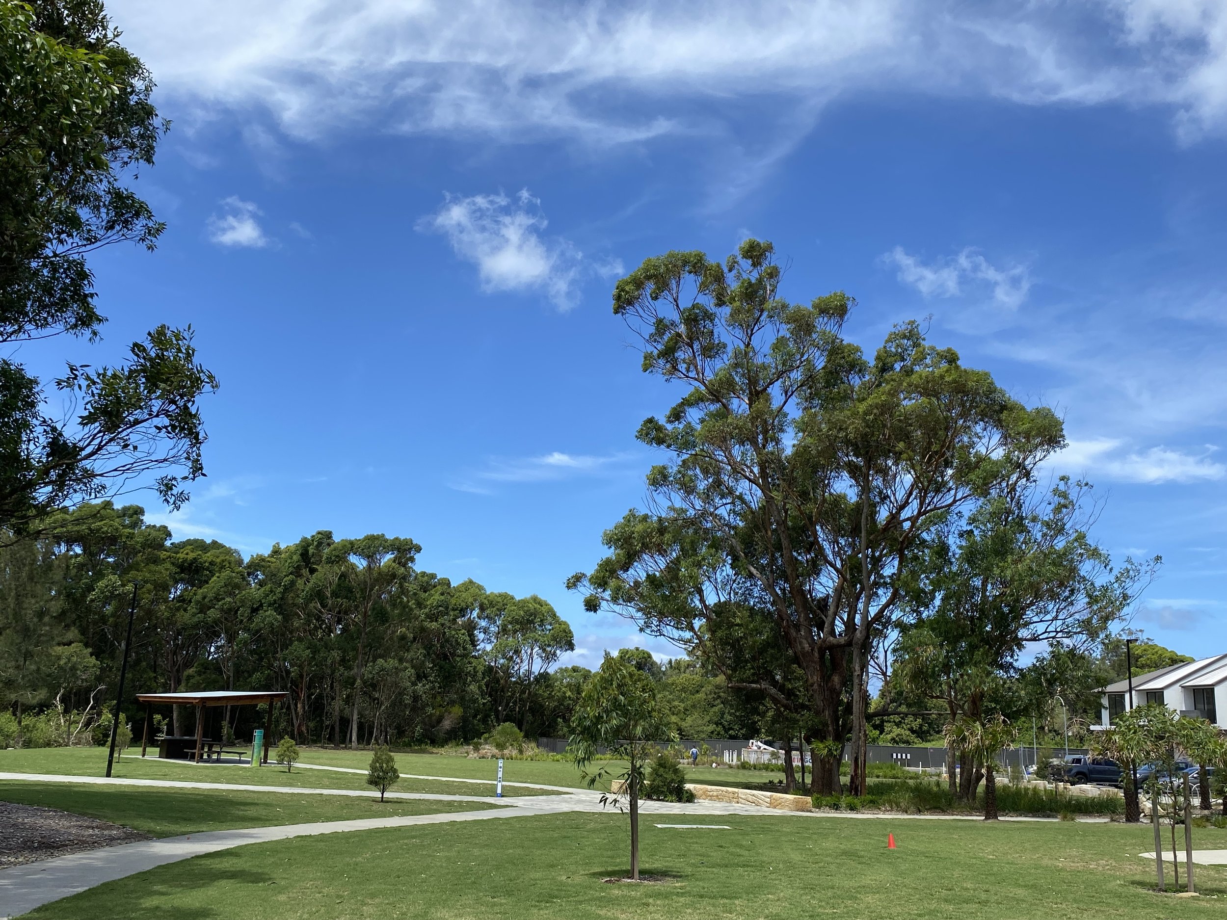 A park with green grass, trees, walking paths, a small shelter, and some buildings in the background under a blue sky with some clouds.
