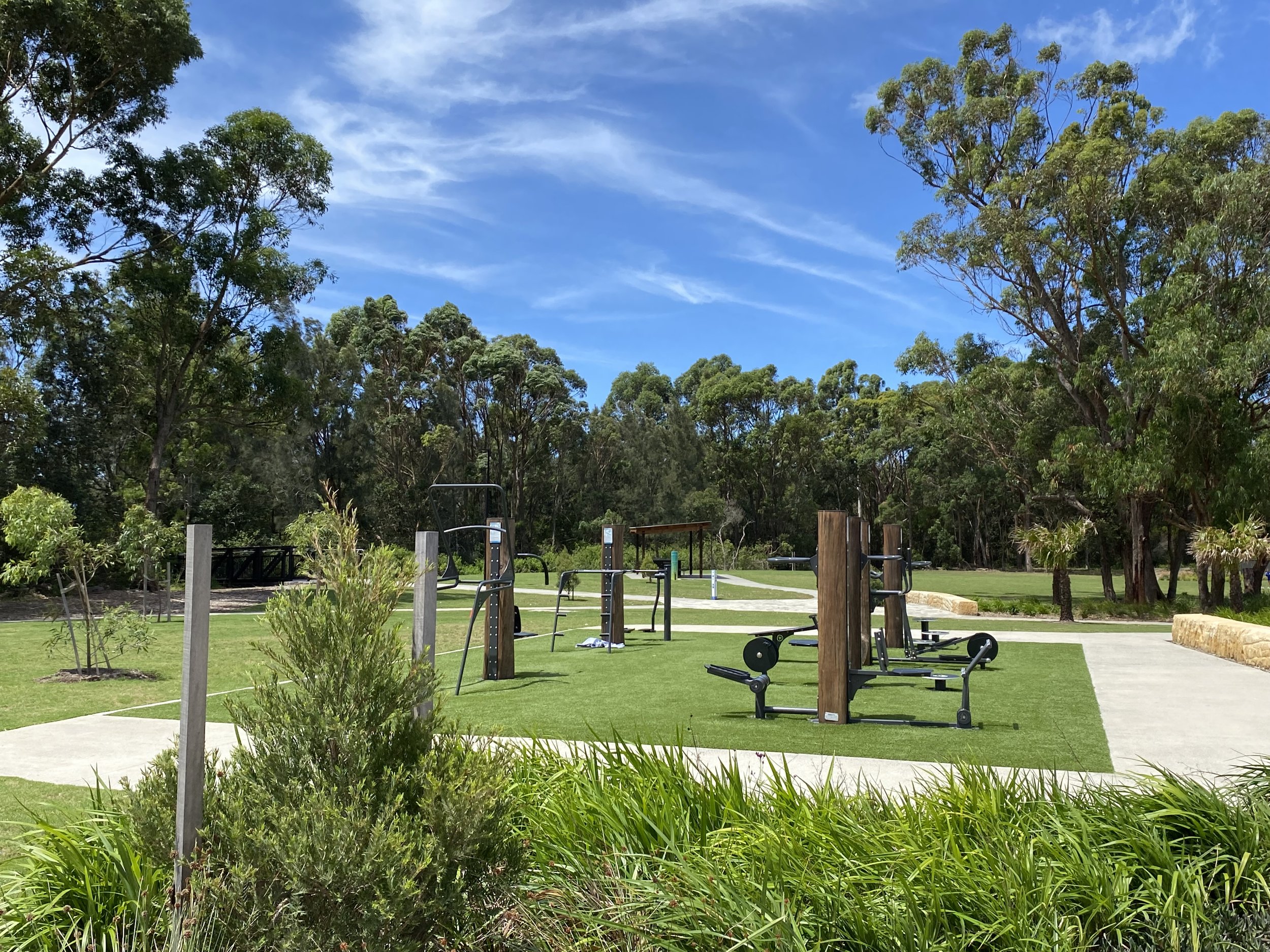 Outdoor fitness park with exercise equipment on artificial grass, surrounded by trees and a clear blue sky.