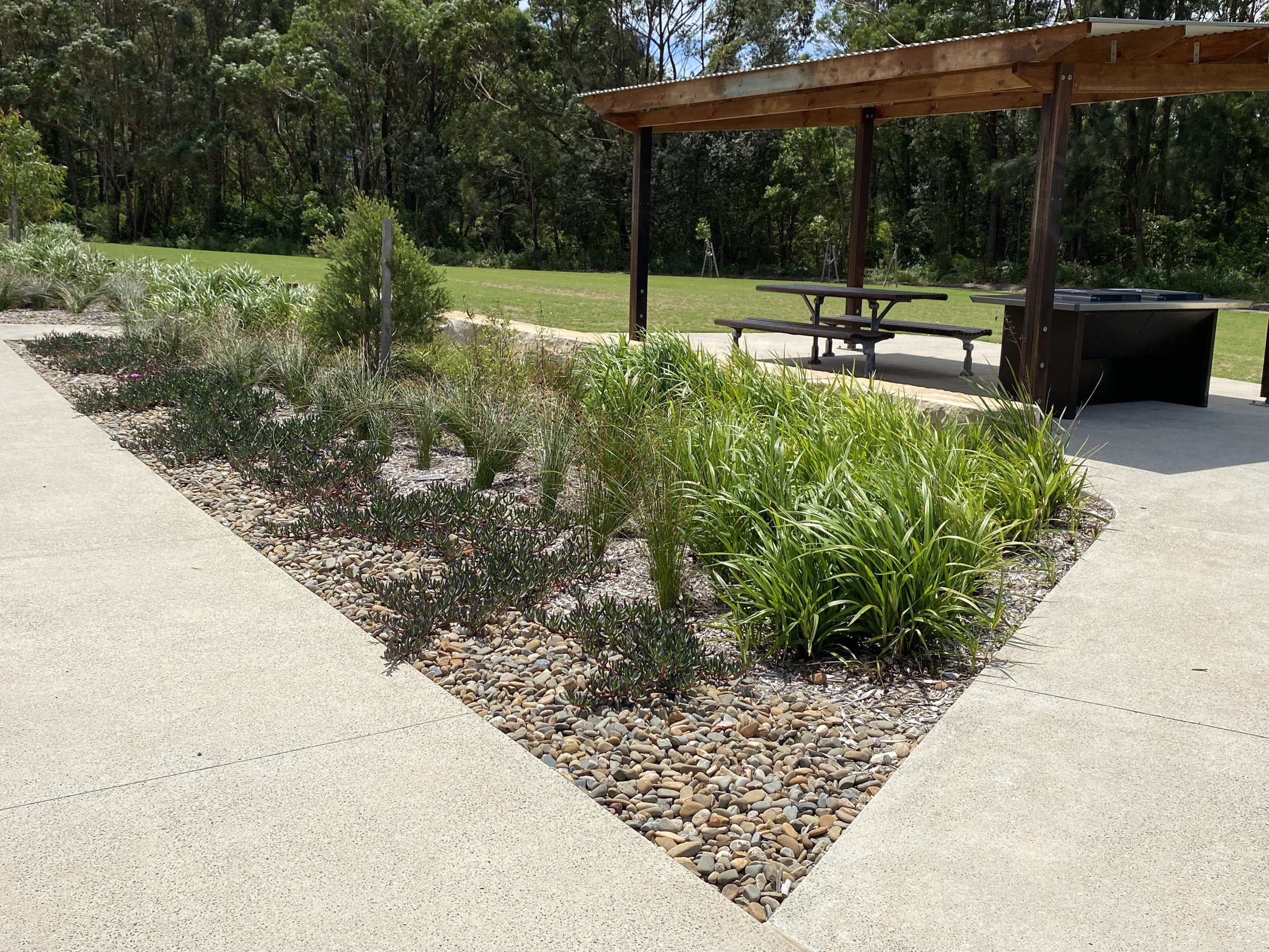 A landscaped park area with a paved walkway, a flower bed with rocks and green plants, a wooden covered picnic area with benches, and a grassy field with trees in the background.