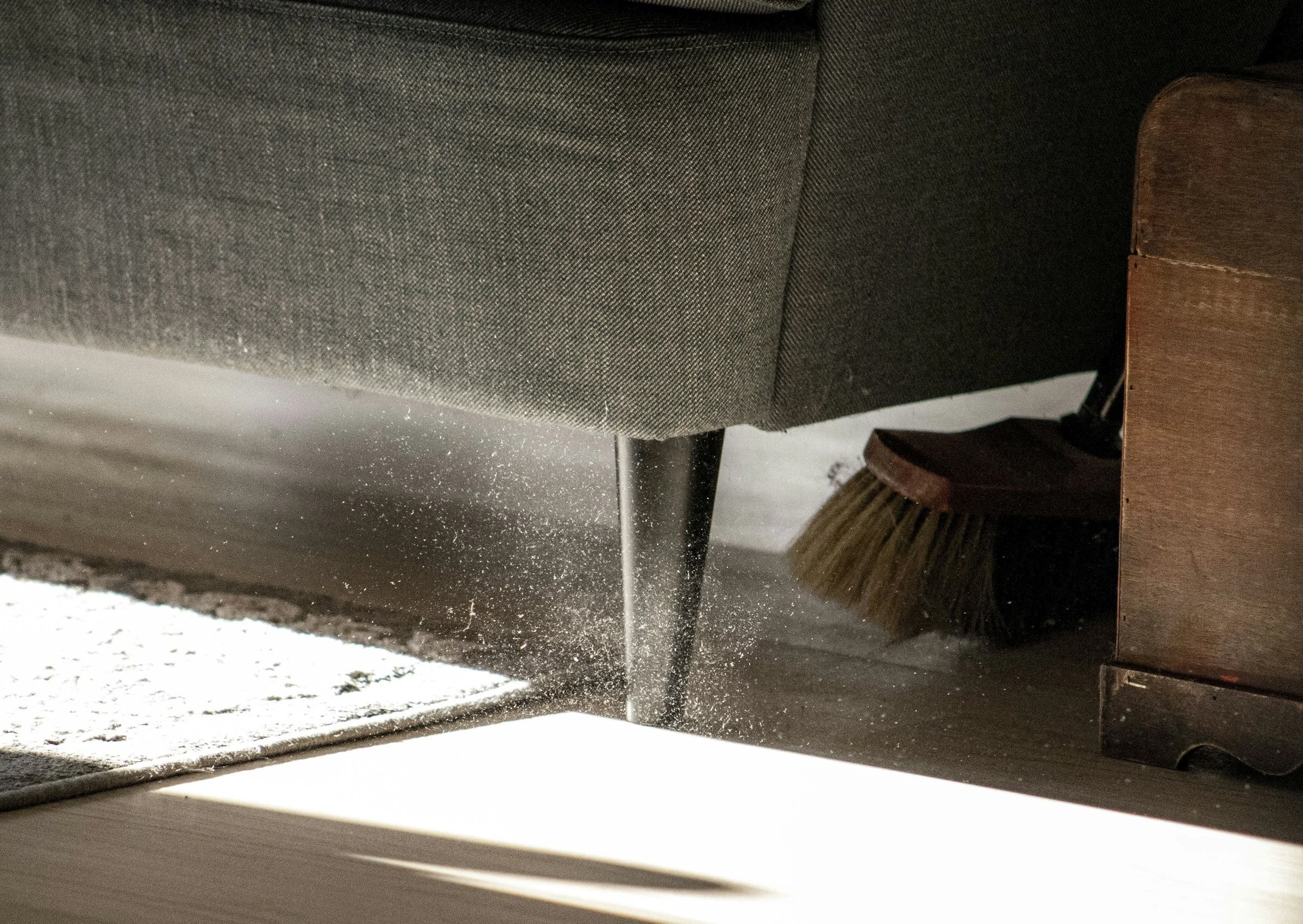 Dust and debris falling from a gap under a sofa onto a floor while a broom and a wooden furniture piece are nearby.
