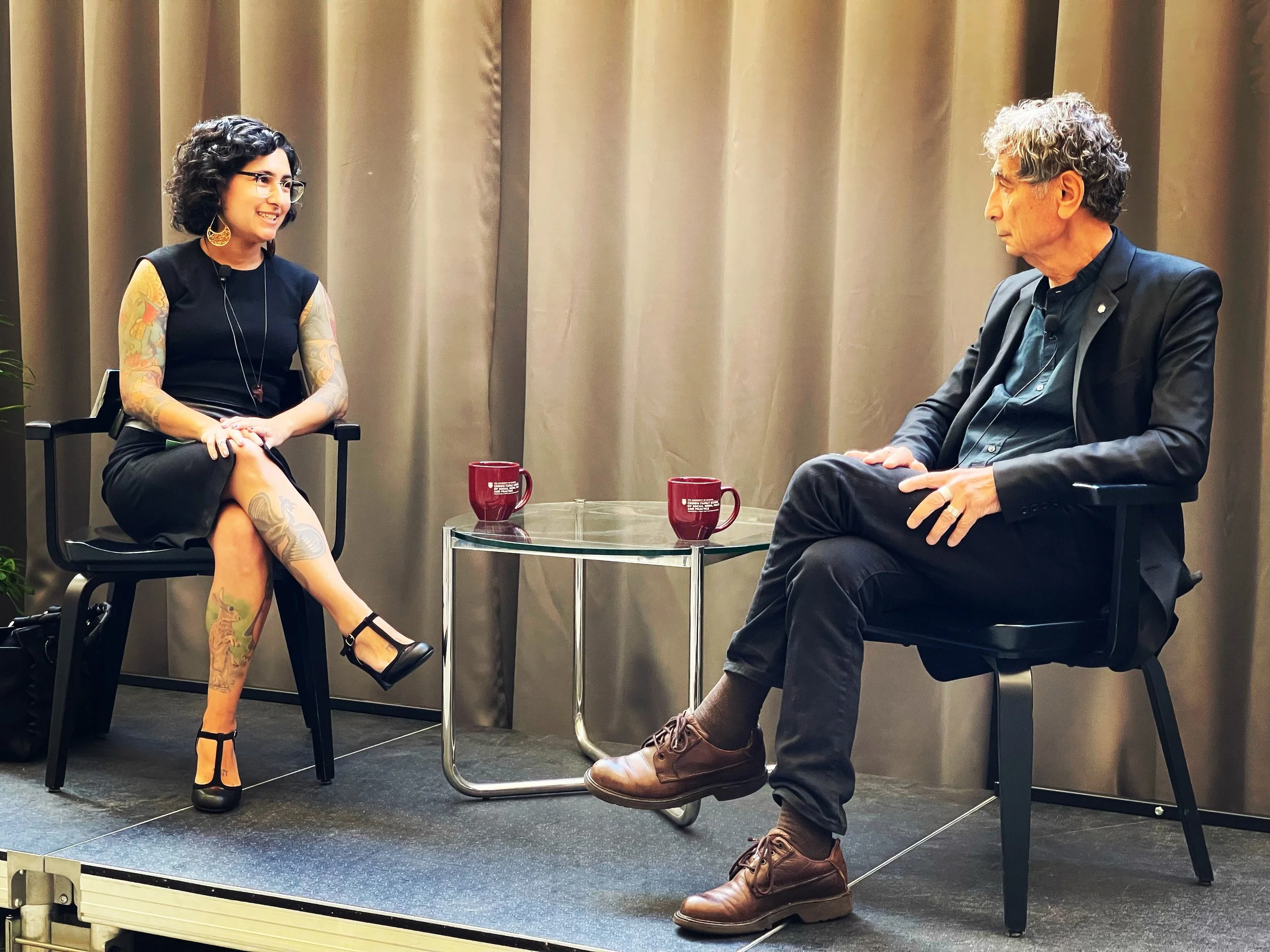Gabriela Zapata-Alma, mental health substance use trauma violence expert, interviews Gabor Mate, trauma and addiction expert, both with wavy black and gray hair and wearing black