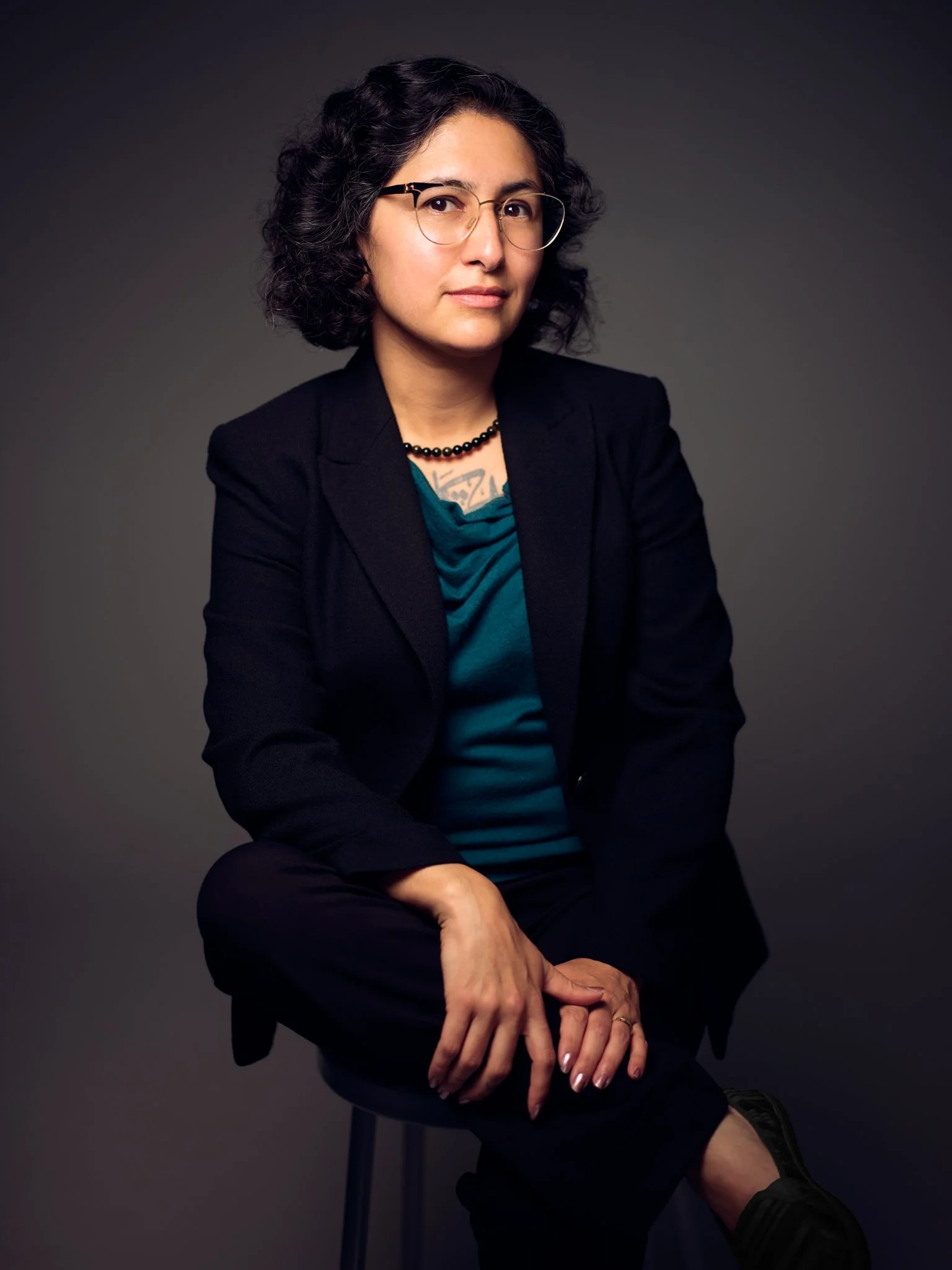 Gabriela Zapata-Alma, licensed clinical social worker and expert trauma therapist, providing compassionate bilingual therapy for trauma and mental health, pictured sitting with glasses, wavy dark hair, a black blazer, teal blouse, and black pants