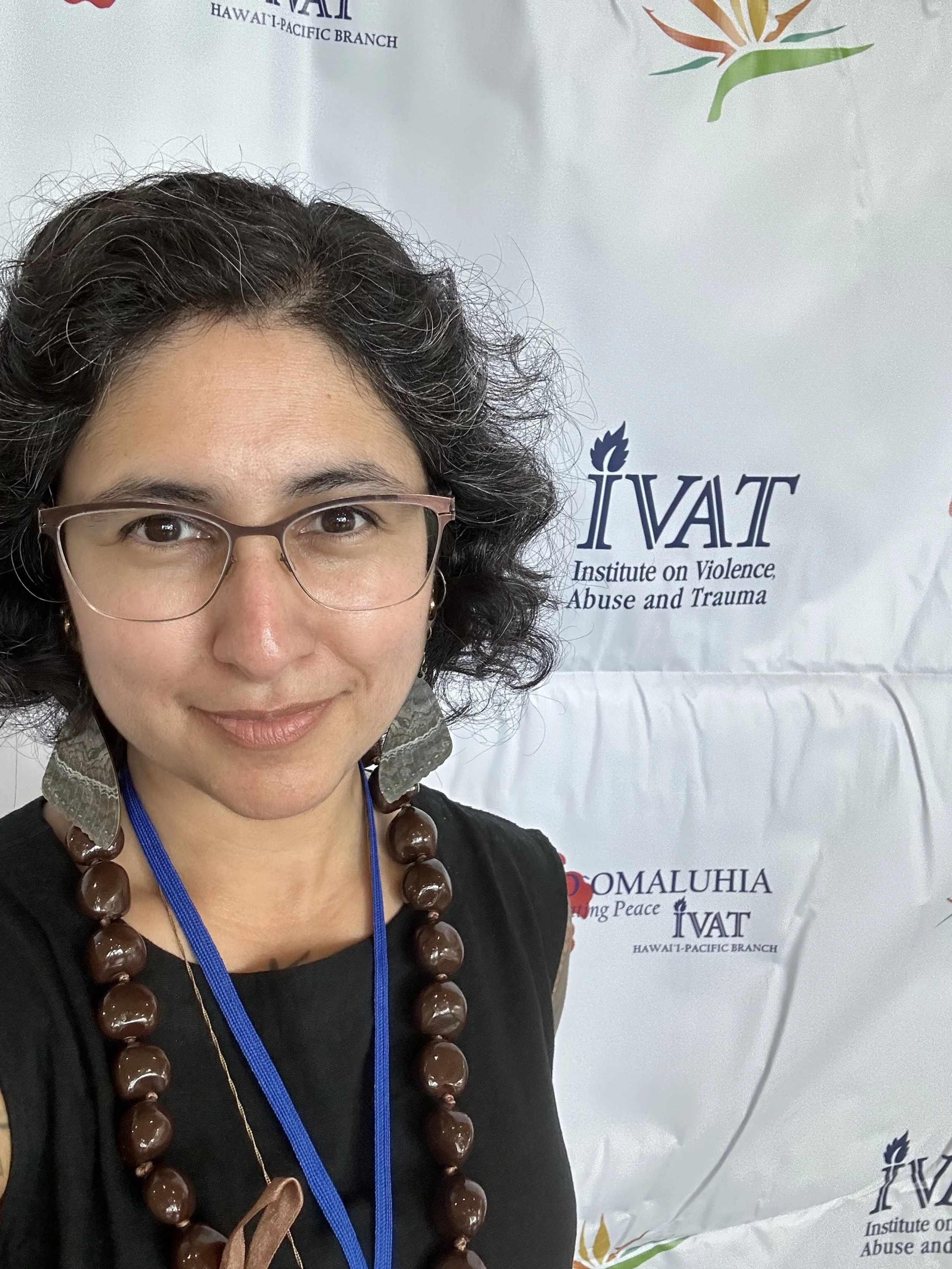 Gabriela Zapata-Alma, Latine with glasses and short wavy hair, trains on domestic violence and substance use coercion at the Institute on Violence, Abuse and Trauma’s Hawaii conference, wearing a black linen top and kukui bead necklace.