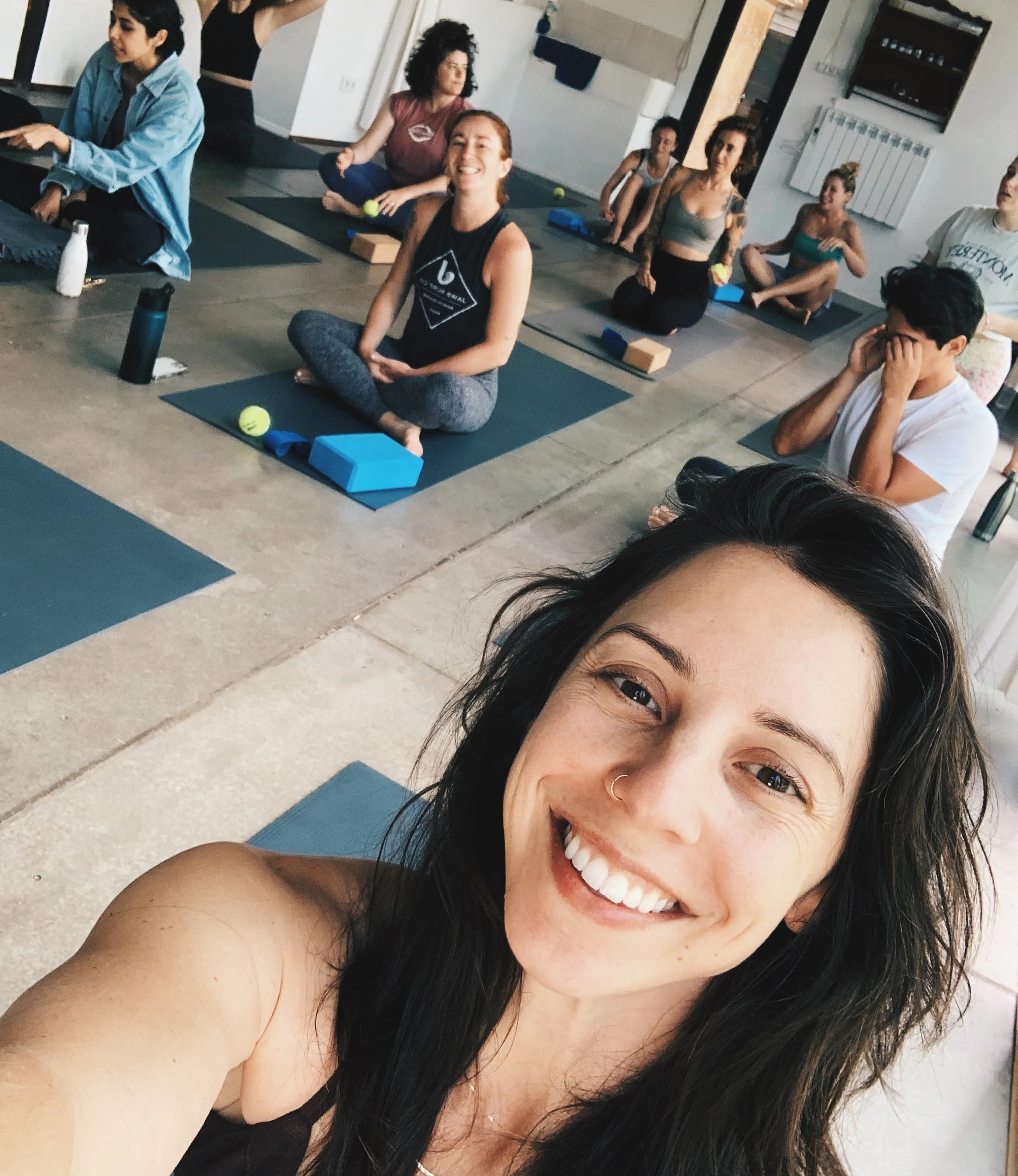 Group of people participating in a yoga or meditation class, sitting on yoga mats with some using blocks and tennis balls, indoors with a facilitator or instructor present.