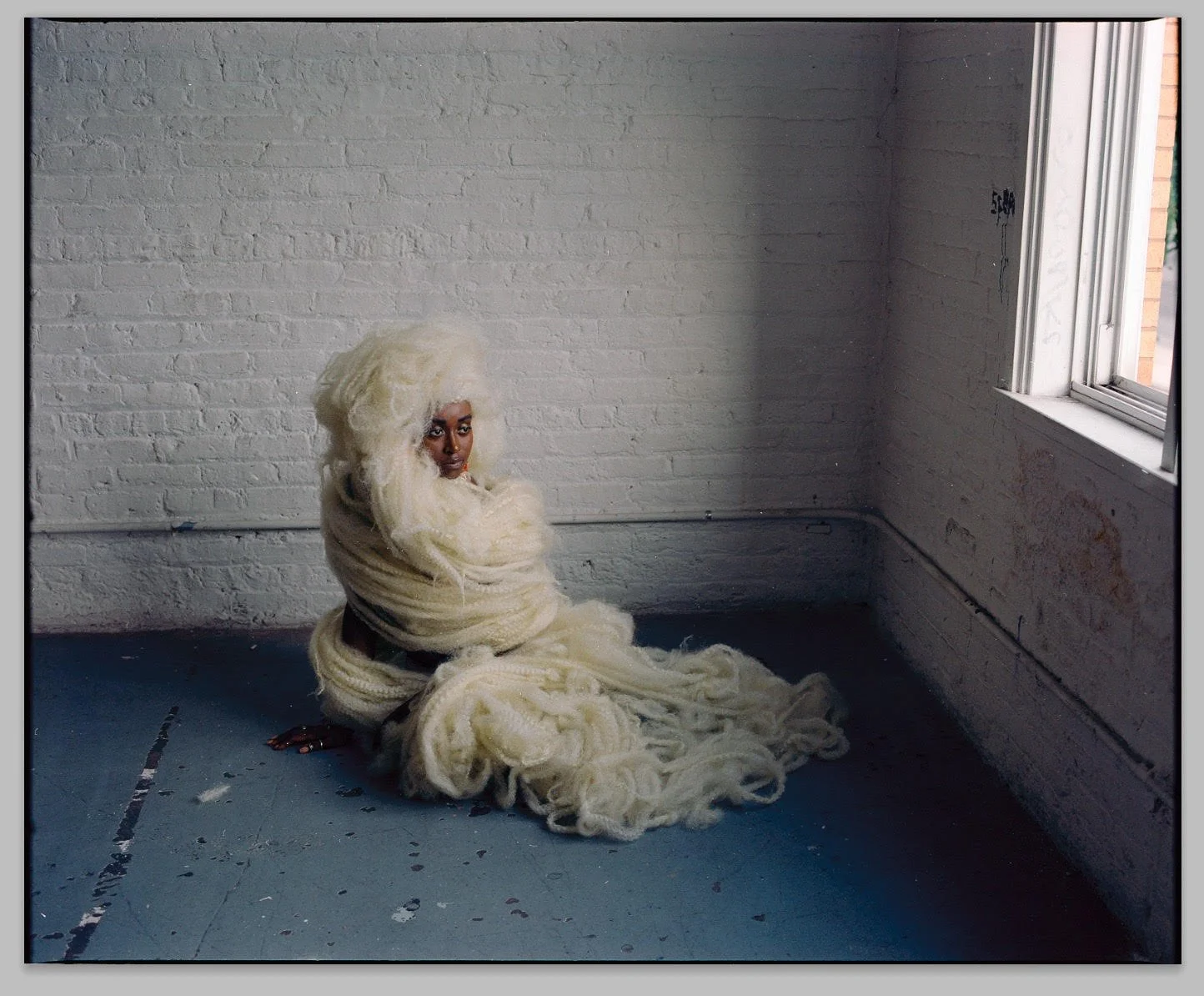 Woman with platinum blonde hair wrapped in large cream-colored yarn, sitting on a gray floor in a minimalist white brick-walled room next to an open window.
