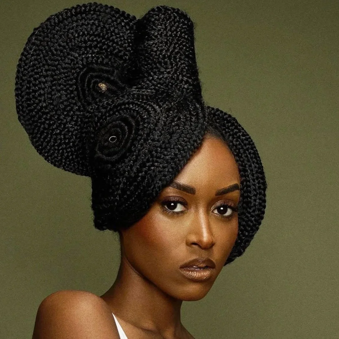 A woman with dark skin features, styled with intricate large black braided hair arranged in a towering updo with spiral details, set against a plain muted background.