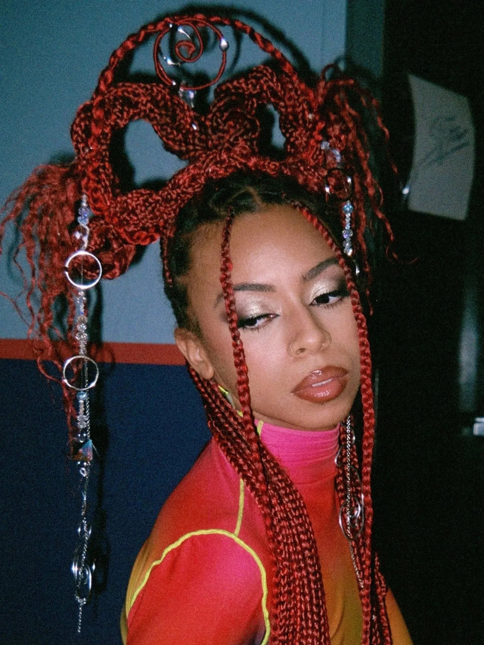 A woman with red braided hair and metallic jewelry poses in front of a dark background. She is wearing a pink and yellow garment.